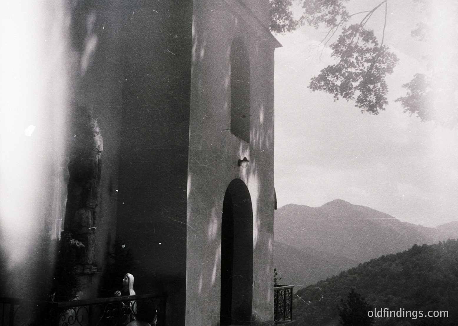Vintage black-and-white photo of a lone figure ascending a stone staircase toward a bell tower entrance, framed by architectural columns. Mountainous landscape and foliage in background suggests elevated, possibly coastal or alpine setting. Mid-20th century architectural style with weathered stonework.