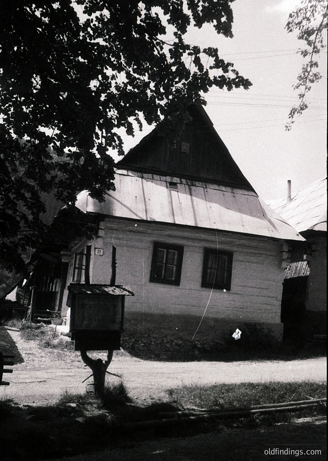 Traditional timber-framed house with steep gabled roof, likely Eastern European rural architecture. Wooden beams and plaster walls suggest 19th–early 20th century construction. Small adjacent structure appears to be a utility shed or storage. Overgrown foliage and cobblestone path hint at historical preservation.