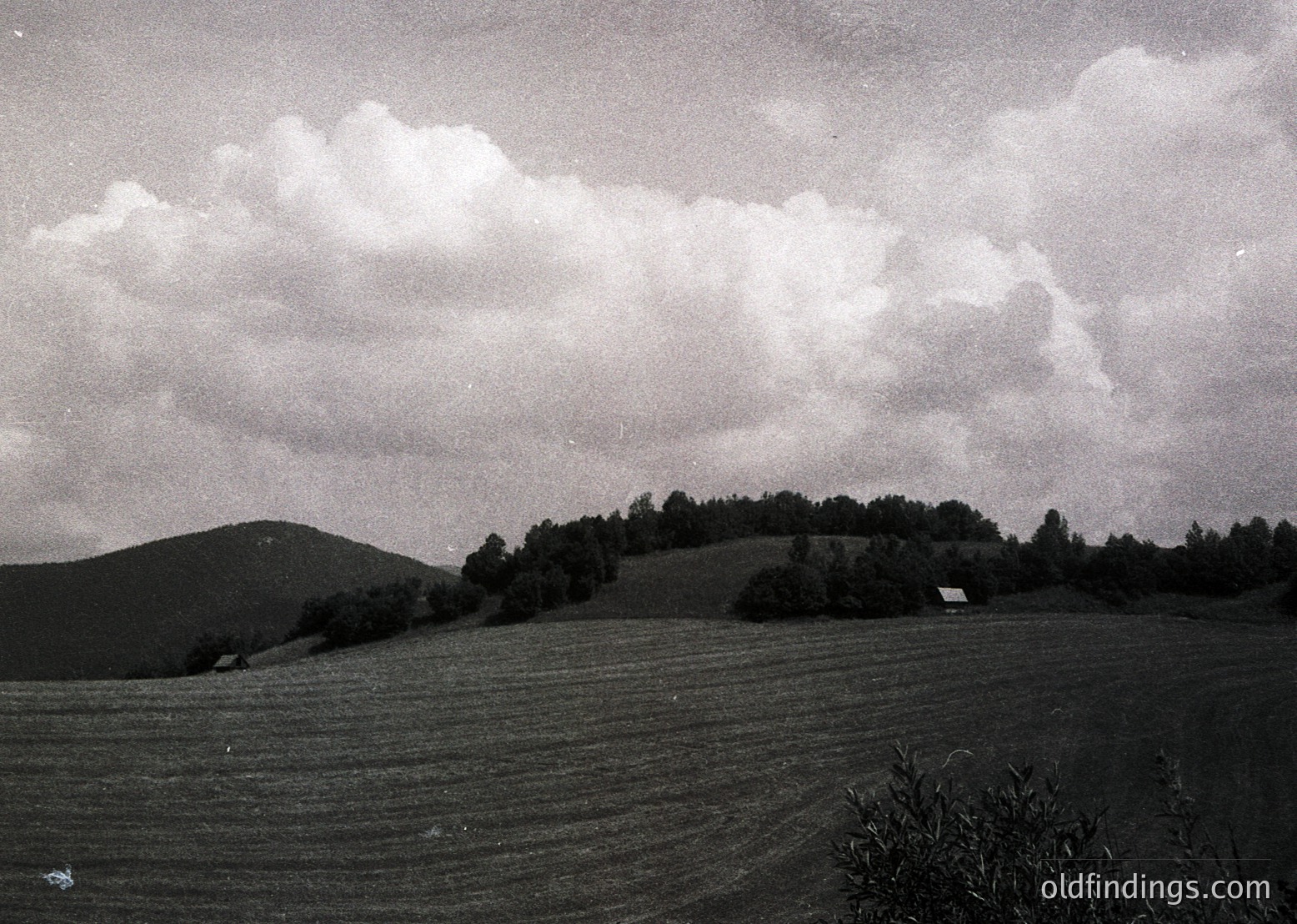 Vintage black-and-white landscape of rolling hills and open fields under dramatic cloud formations. A lone farmhouse sits near treeline, suggesting rural isolation. Grainy texture and soft focus evoke mid-20th century photography. Ideal for historical or nostalgic design references.