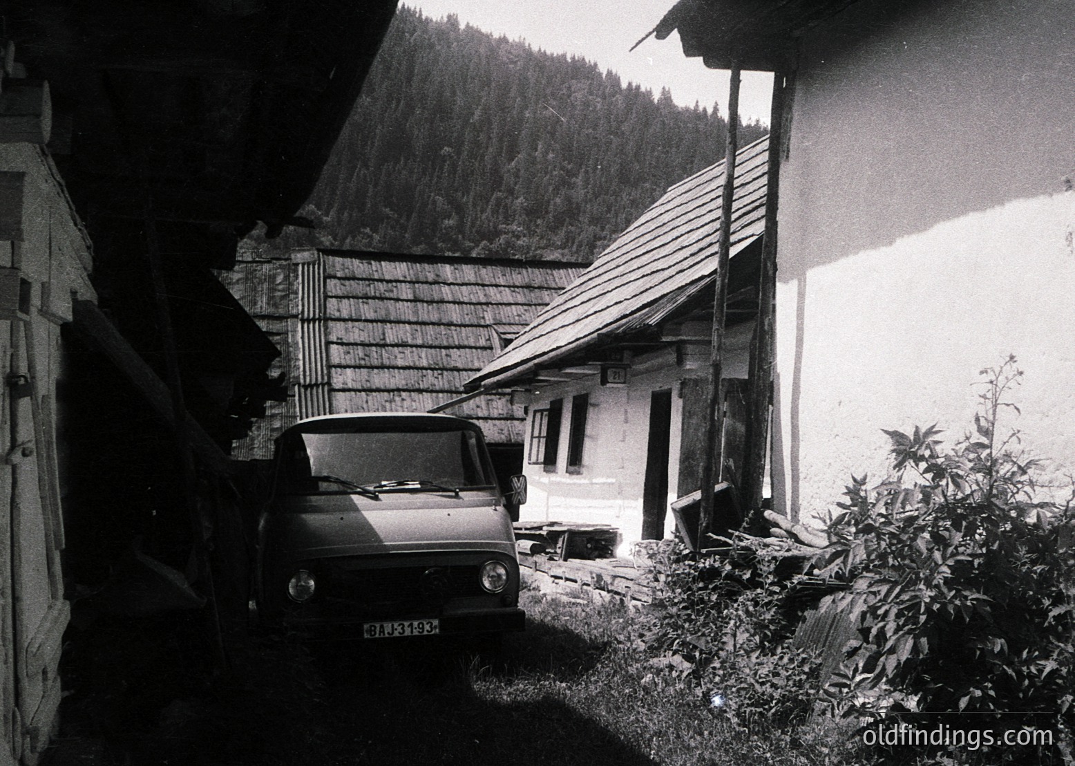 Classic mid-century Alpine cottage with rustic stonework and wooden shingle roof, nestled in a forested valley. A vintage van (likely 1960s–70s) parked beside a narrow pathway, framed by overgrown greenery.