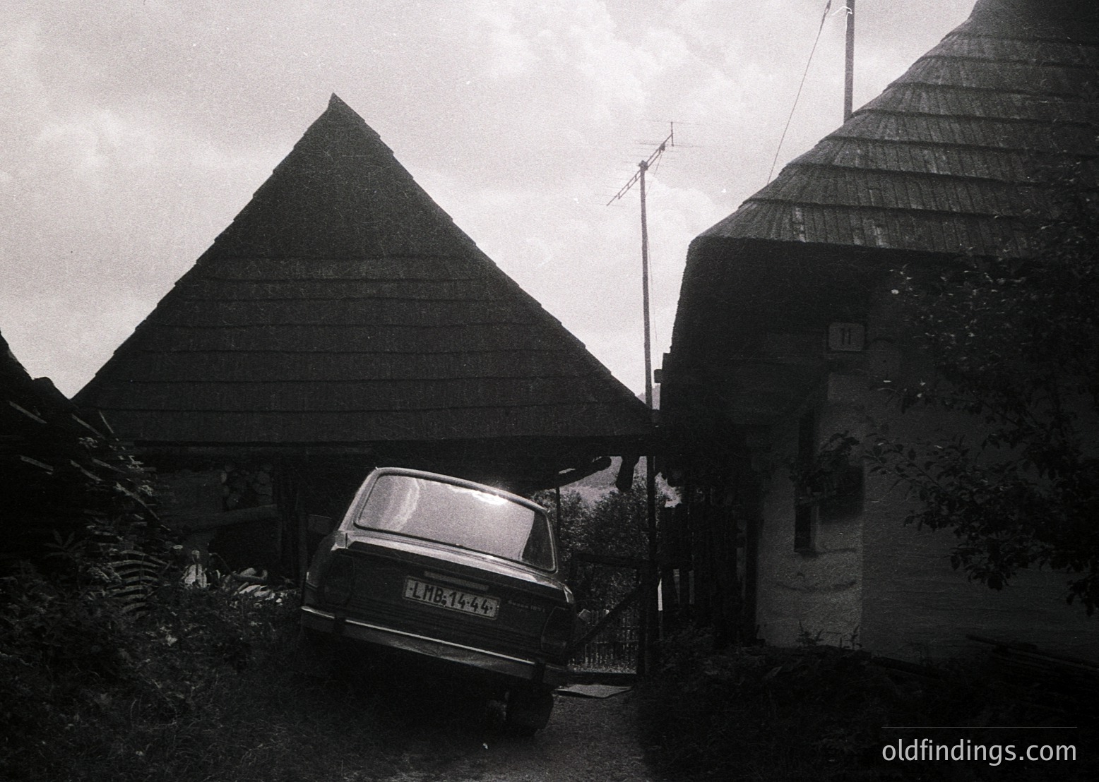 Black-and-white rural scene featuring two distinct traditional structures: a conical wooden barn and a cylindrical hayloft. A vintage car (likely 1960s–1980s) is parked under a covered entryway. Overgrown greenery and a rustic fence frame the composition. Evokes Eastern European countryside architecture.