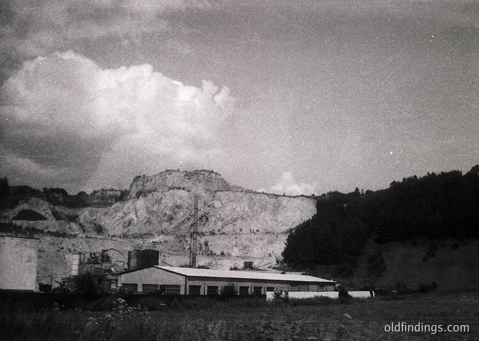 Mid-century industrial landscape featuring a low-slung, flat-roofed building beside a rocky quarry face. Dramatic cloud formations contrast with the rugged terrain, suggesting a post-war or early industrial era. Likely Eastern European due to architectural style.