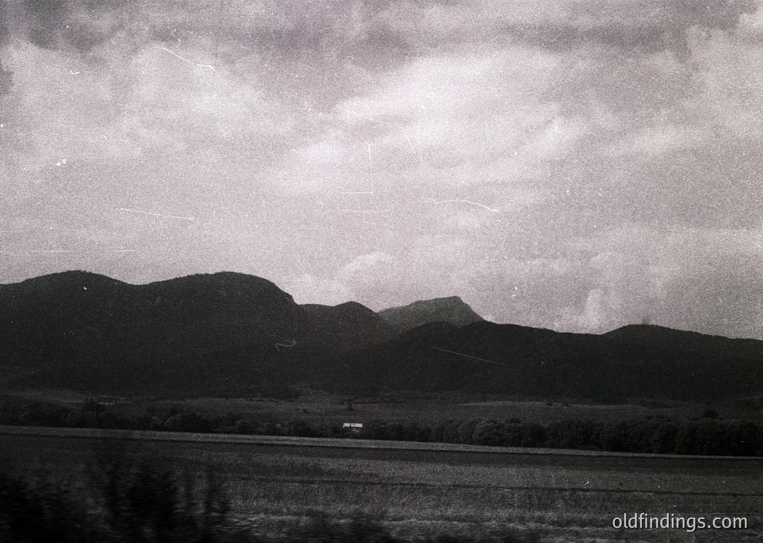 Mid-20th century black-and-white landscape featuring rolling hills, dense cloud cover, and a lone road winding through flat terrain. Reflections in a body of water below suggest a serene, possibly rural setting. Vintage aesthetic captures atmospheric drama.