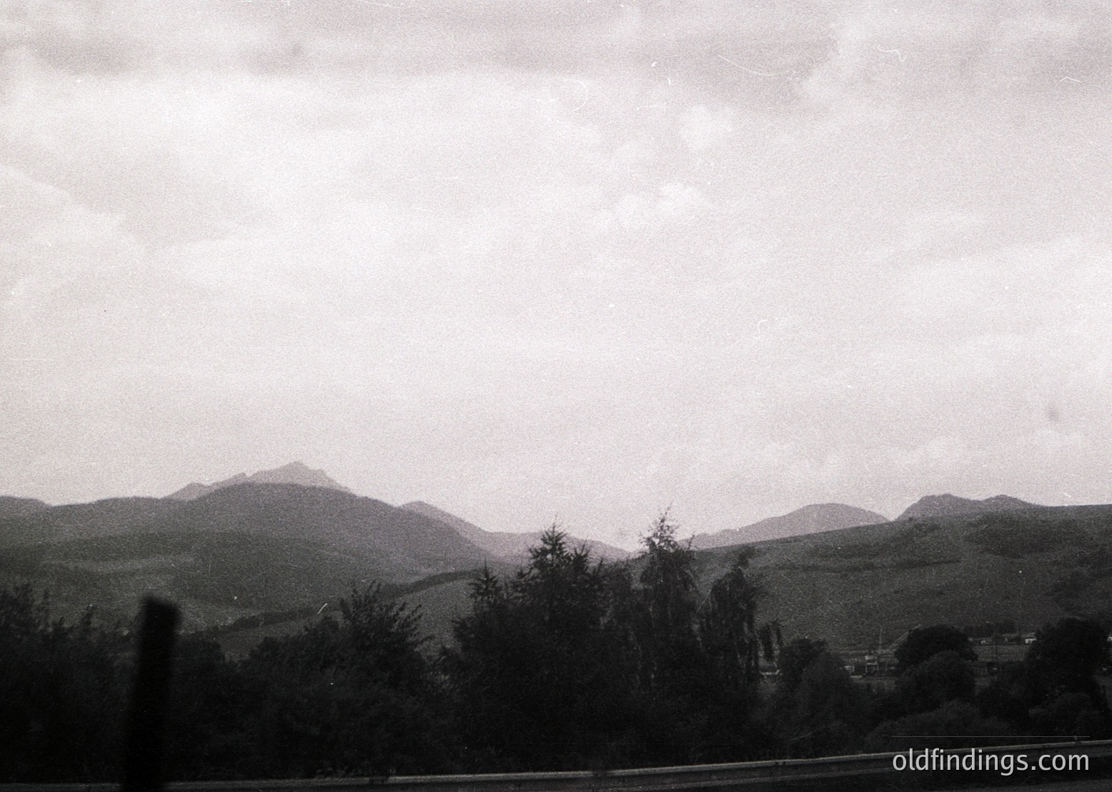 Vintage black-and-white landscape featuring rolling hills and misty mountains under overcast skies. Foreground shows dense tree line, likely coniferous, with subtle road or path visible. Atmospheric, moody lighting suggests early photography era.