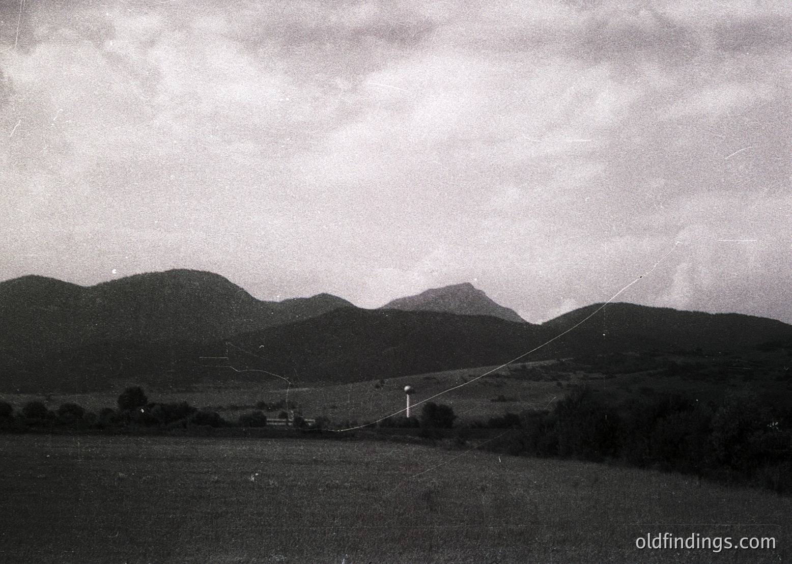 Black-and-white landscape featuring rolling hills and a lone utility pole with power lines stretching diagonally across the frame. Low-contrast skies suggest overcast conditions. Mid-20th century rural setting, likely European.