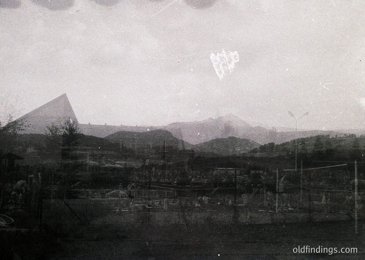 Vintage black-and-white aerial view of industrial ruins and mountainous terrain. Overgrown machinery and skeletal structures hint at post-war abandonment. Distinctive pyramid-shaped industrial chimney dominates skyline. Likely Eastern European, mid-20th century.