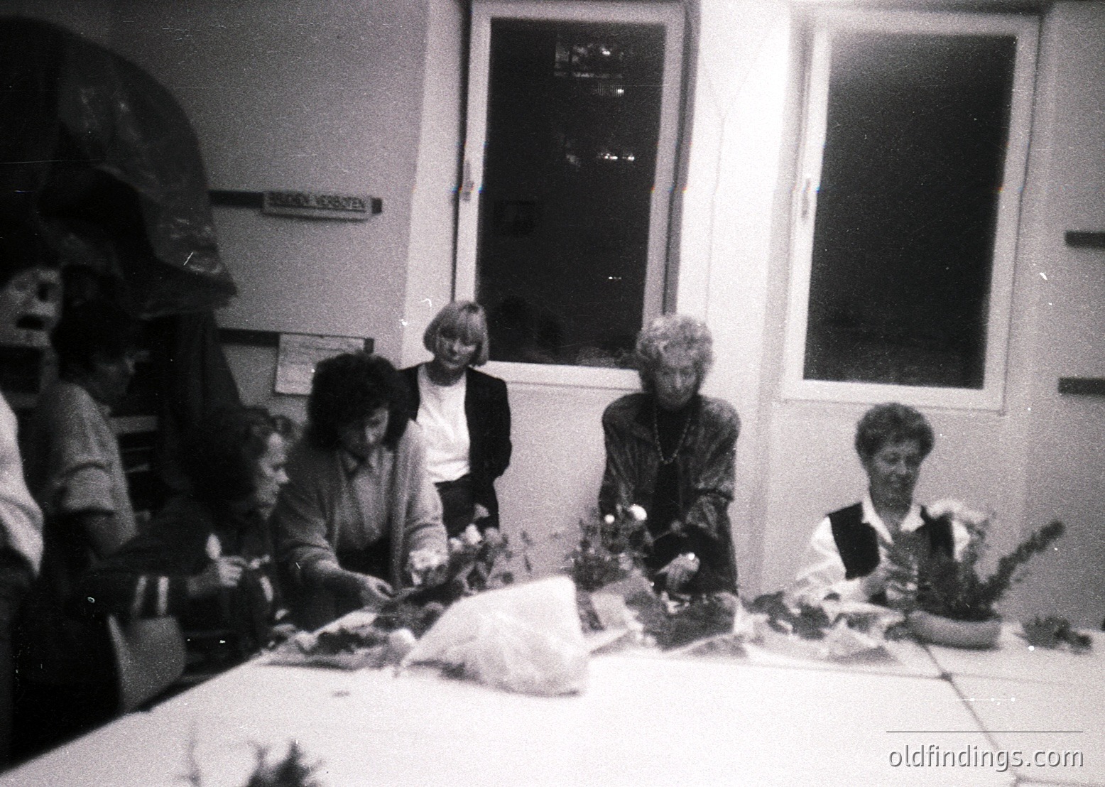 Vintage black-and-white photo of a group of women gathered around a table, crafting floral arrangements. Indoor setting with a tiled floor and white-painted walls. Sign on wall reads "БЕЛГРАДСКА ЗЕМЈА" (Belgrade Region). Mid-20th century clothing and floral styles suggest or .