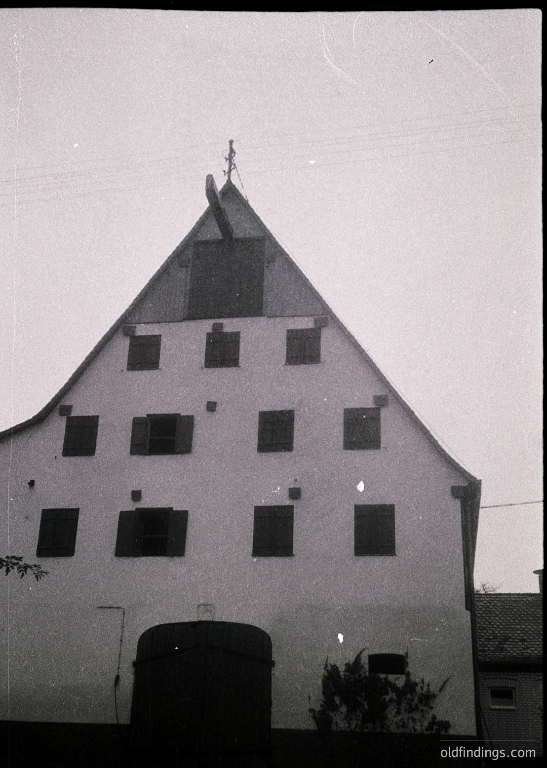 Three-story gabled farmhouse with steep, symmetrical roof and small rectangular windows. Central arched entrance with weathered stone trim. Cross finial atop gable peak. Likely 19th-century rural European architecture.