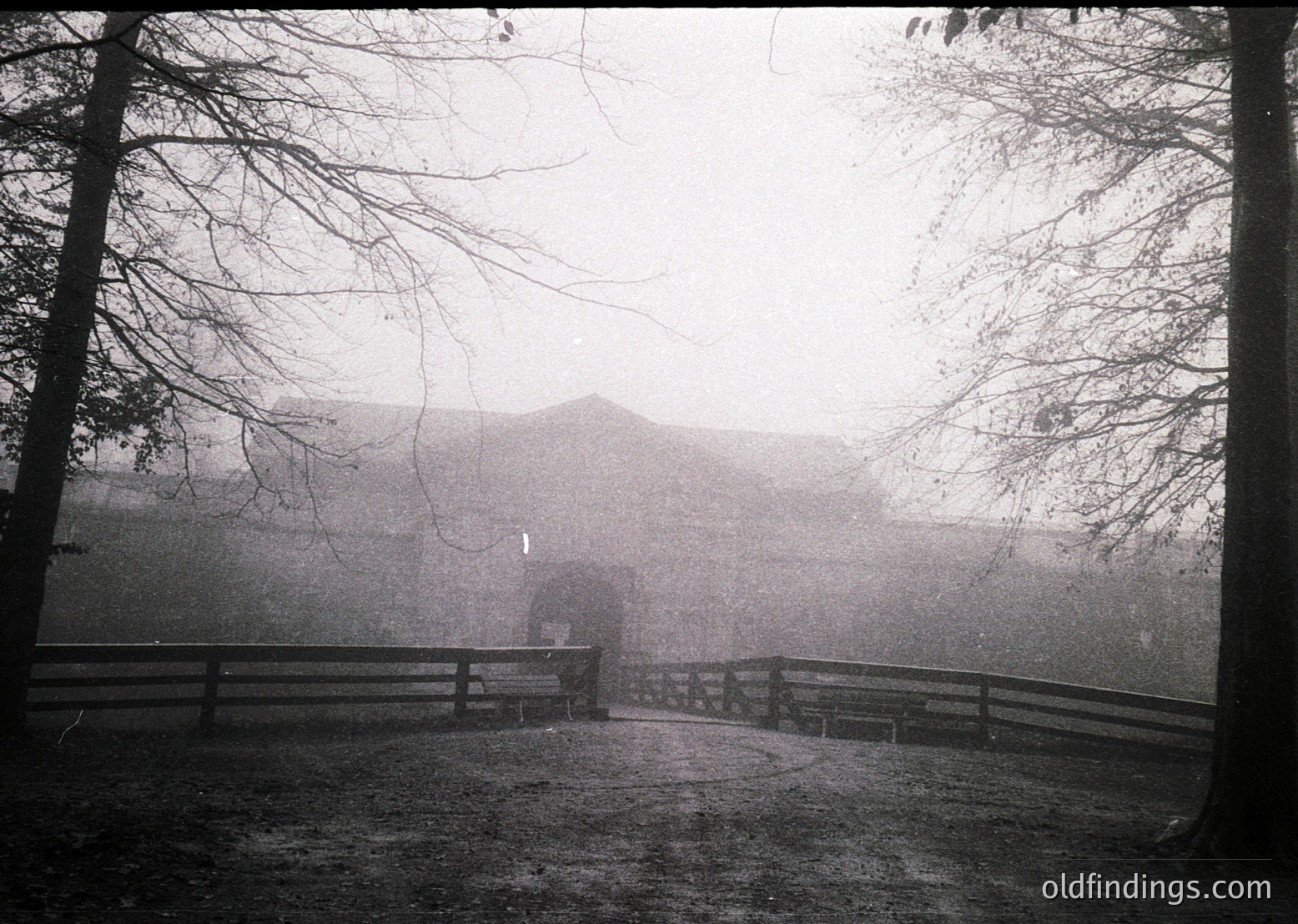 Misty black-and-white landscape featuring a fog-shrouded pathway flanked by wooden fencing, leading to a distant, indistinct building. Bare trees frame the scene, enhancing atmospheric depth. Likely early-to-mid 20th century rural or estate setting.