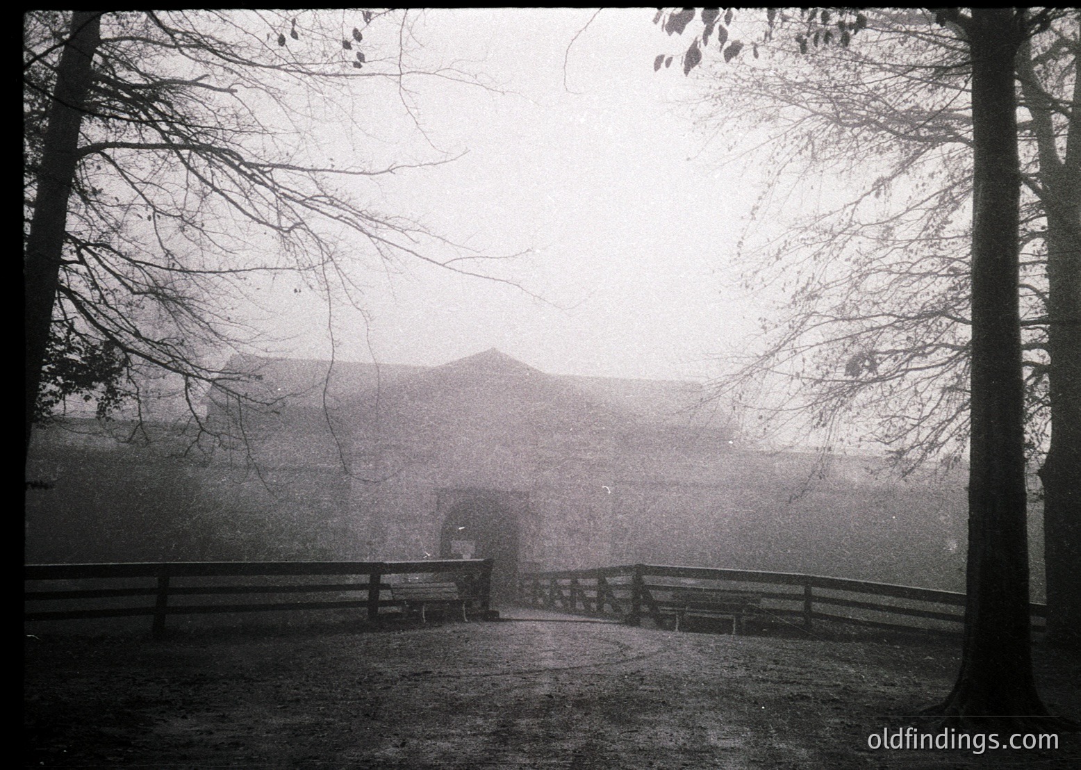 Vintage black-and-white photo of a misty pathway flanked by wooden railings, leading to a distant, partially obscured building. Bare trees frame the scene, suggesting late autumn/winter. Fog obscures details, creating an atmospheric, moody composition. Likely early-to-mid 20th century.