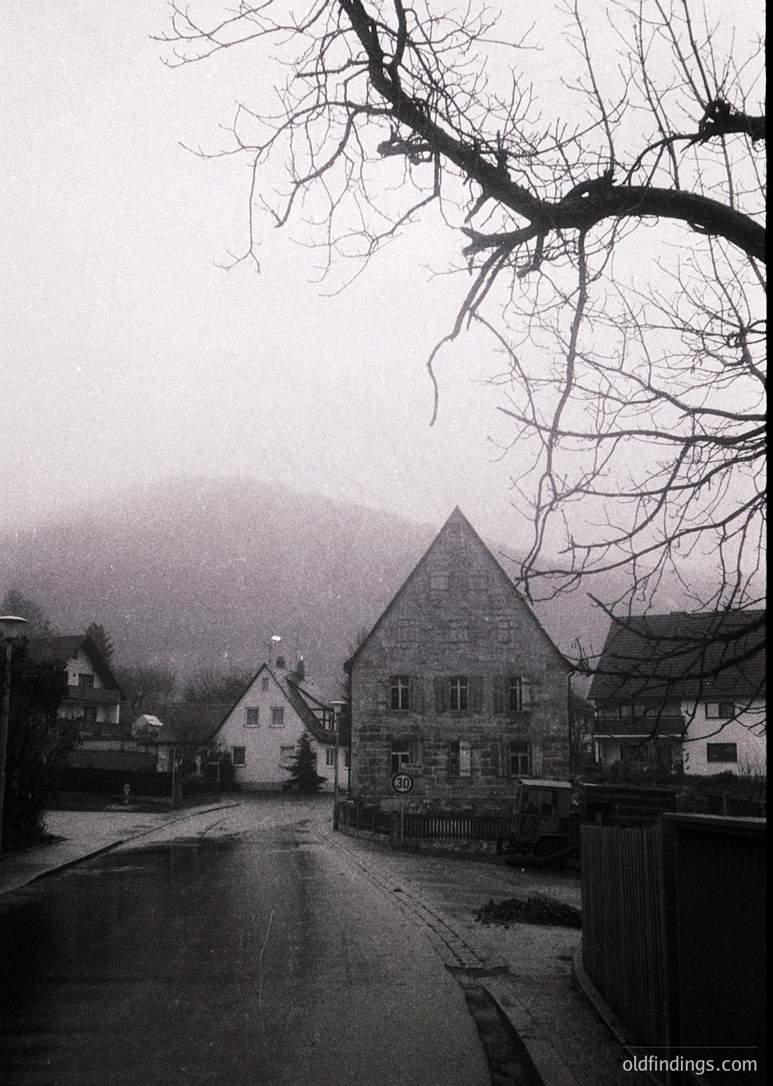 Misty village street with stone buildings, including a prominent gabled structure and smaller half-timbered homes. Bare trees frame the scene, suggesting late autumn/winter. Fog obscures distant hills, enhancing atmospheric depth. Likely Alpine or Central European rural setting.