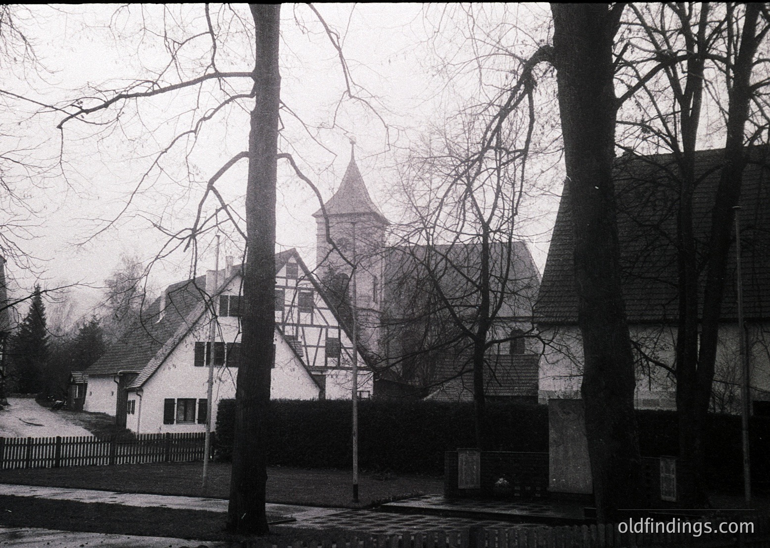 Black-and-white shot of a **half-timbered Tudor-style church** with a steep gable roof and central tower, framed by bare winter trees. Surrounding residential buildings exhibit similar architectural style, suggesting a historic European village. Foggy, muted lighting enhances the nostalgic atmosphere. Likely **1950s–1970s** based on architectural details and photographic style.