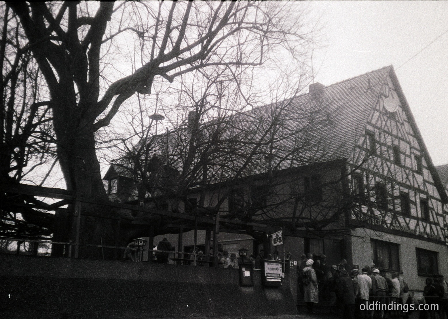 Timber-framed half-timbered building with exposed beams, likely a traditional European inn or restaurant. Crowd of people gathered outside, suggesting a social or public event. Leafless trees indicate late autumn/winter. Black-and-white photo suggests 1950s–1970s era.