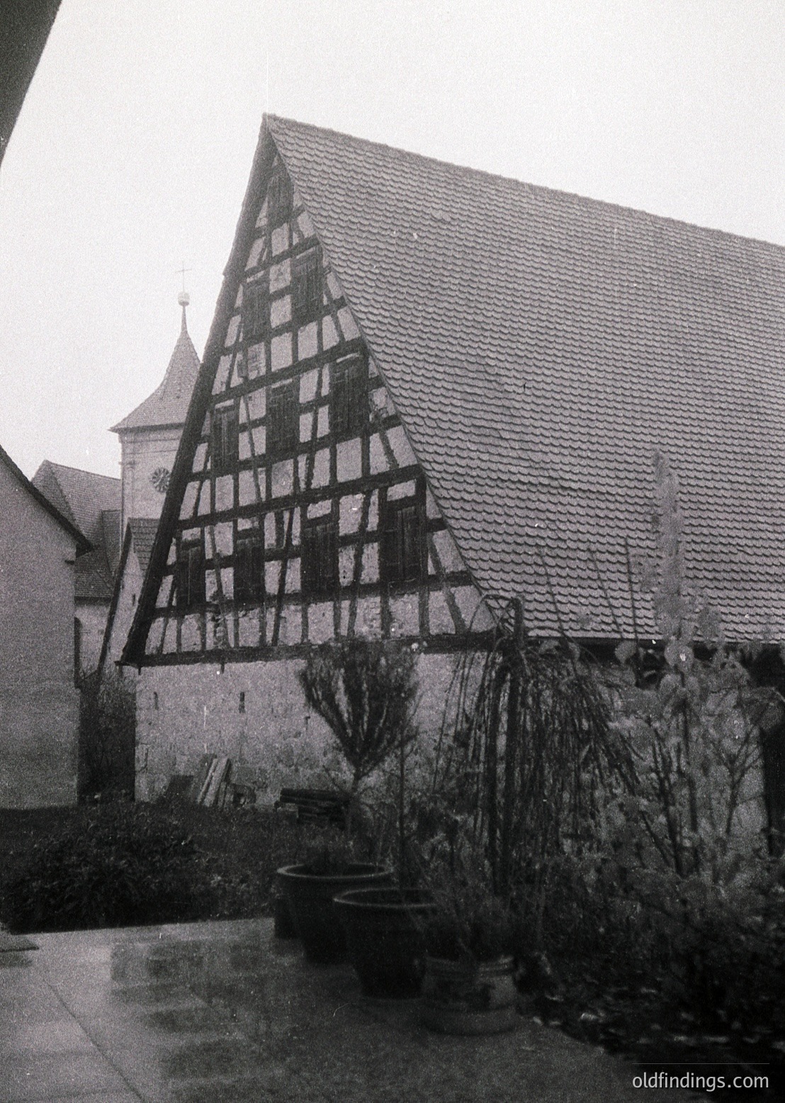 Timber-framed half-timbered house with exposed black-and-white weave, likely German Alpine or Black Forest architecture. Courtyard features potted plants and a stone pathway. Overcast sky enhances moody, vintage atmosphere.