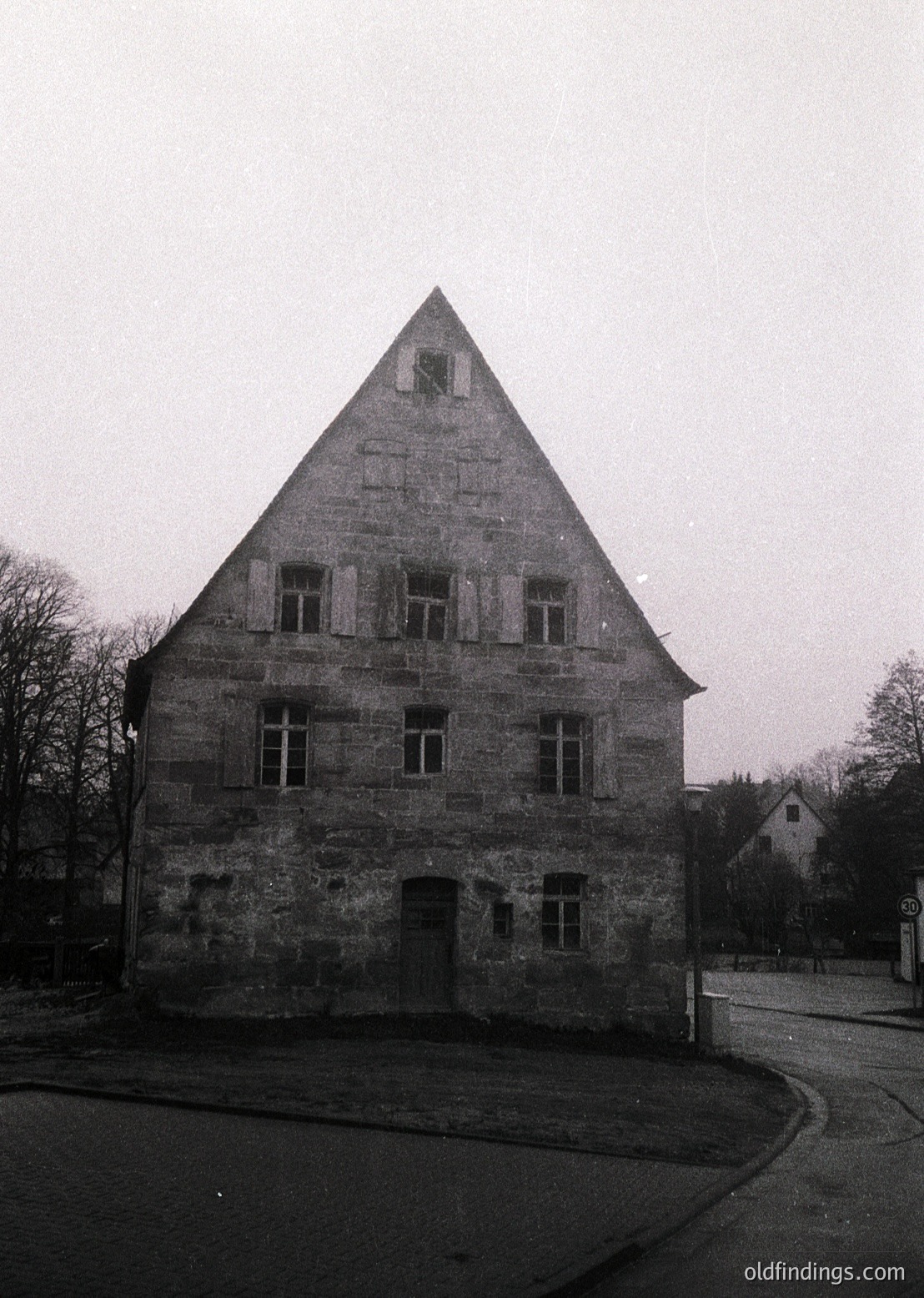 Historic stone building with steep gabled roof, likely 18th–19th century European architecture. Symmetrical facade features three tall windows on upper level and a central arched entrance below. Surrounding bare trees suggest late autumn/winter. Urban setting with paved road and minimal modern interference.
