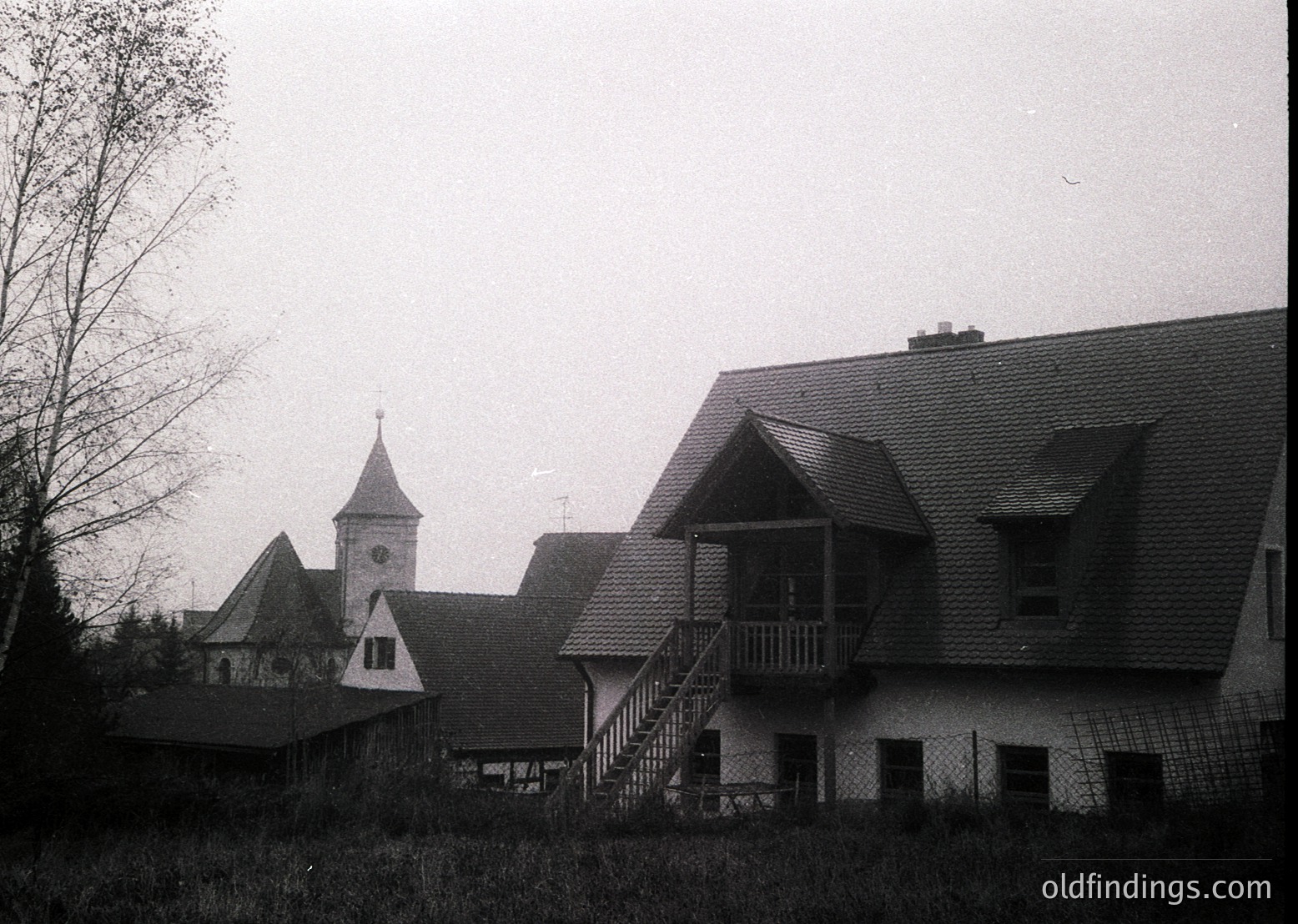 Black-and-white photo of a mid-20th century European village. Prominent gabled roofs, brick chimneys, and a church steeple with a cross dominate the scene. Wooden balconies and laundry lines suggest residential use. Overgrown grass and leafless trees indicate late autumn/winter. Likely Eastern European,