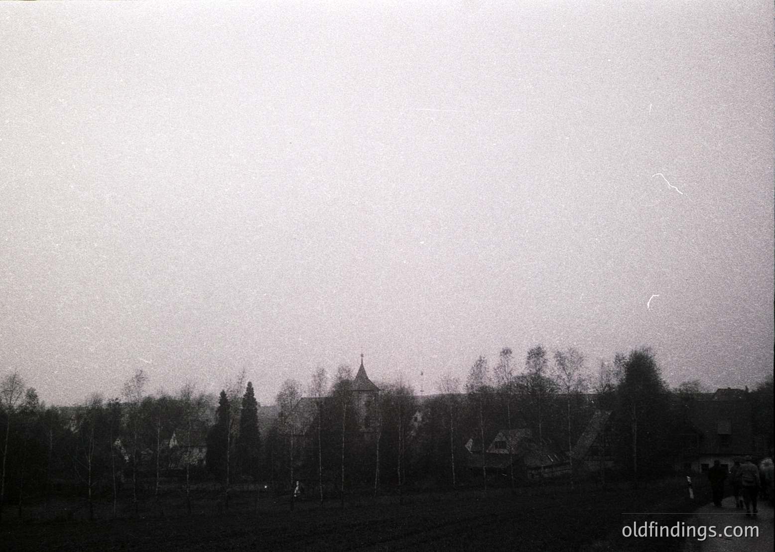 Vintage black-and-white shot of a rural cemetery with tall, uniform gravestones and a central chapel. Leafless trees frame the scene, suggesting late autumn or winter. Overcast sky enhances the solemn atmosphere. Likely Eastern European, mid-20th century.