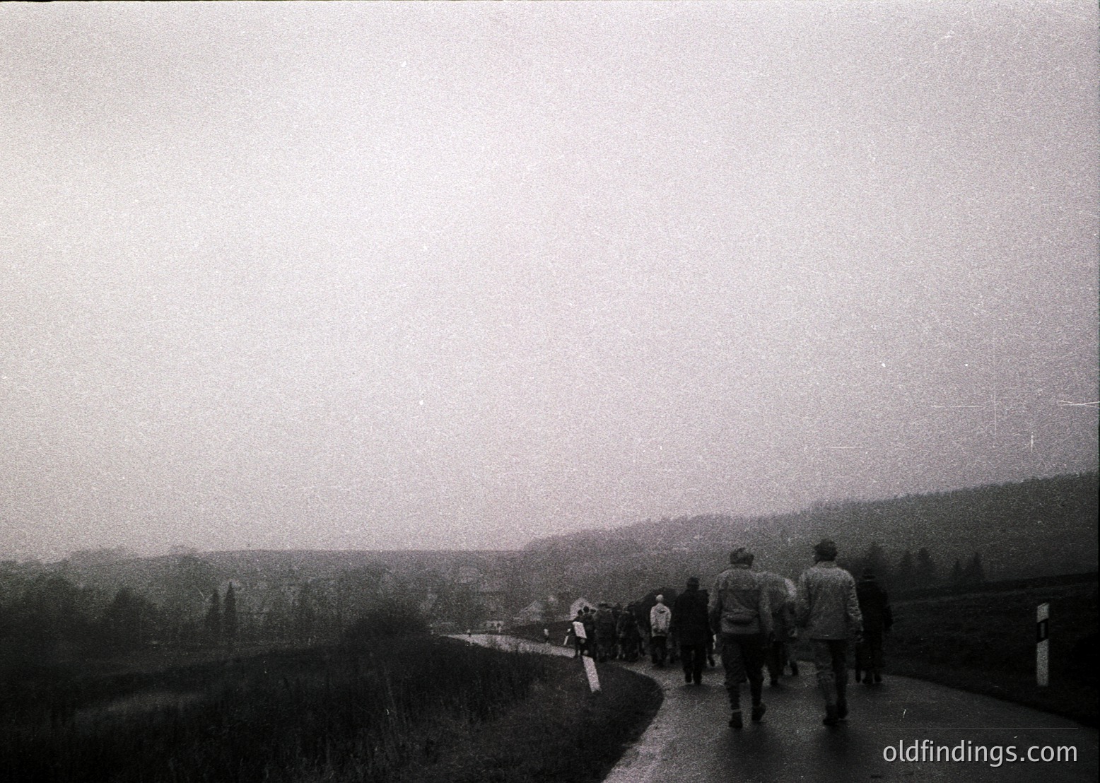 Black-and-white street scene featuring a group of 12+ pedestrians walking along a paved road flanked by grassy areas and distant hills. Foggy, overcast conditions obscure visibility. Clothing suggests mid-20th century (1950s–1970s) style.
