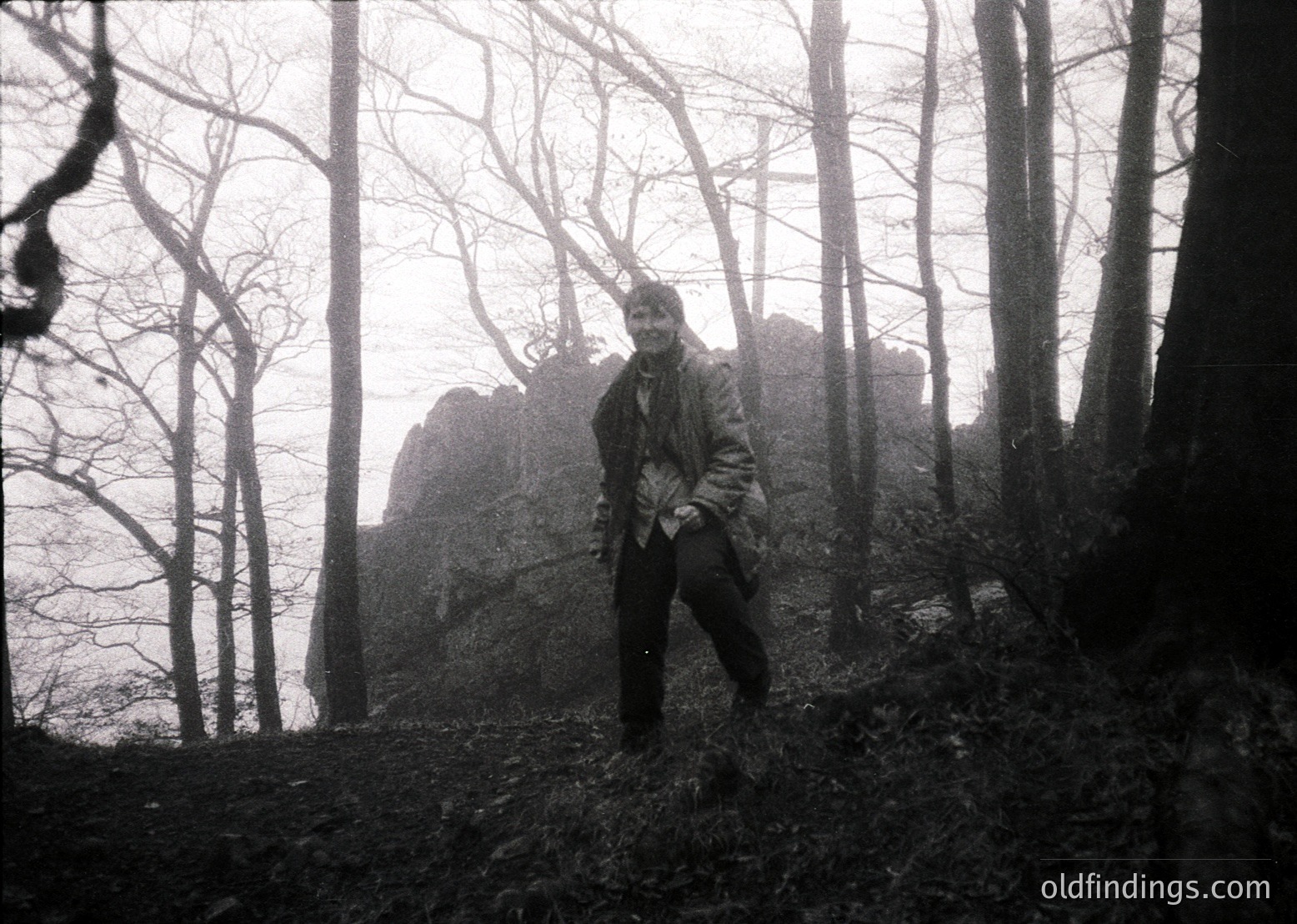 Black-and-white portrait of a lone individual in a dense forest, standing near a rocky outcrop. Clothing suggests mid-20th century outdoor attire. Misty, overcast atmosphere enhances dramatic mood. Likely European alpine or temperate forest setting.