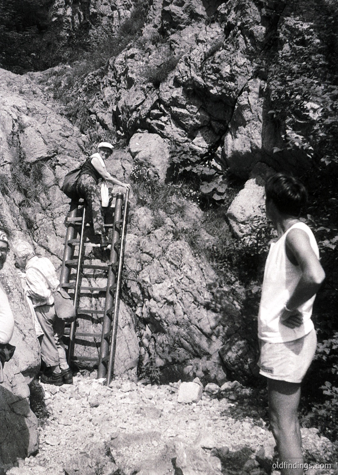 Three individuals navigate a rugged, rocky terrain using a metal ladder in mid-20th century attire. The scene suggests construction or excavation work, likely in a mountainous or hilly region. The person on the ladder wears a wide-brimmed hat and shorts, while the others in shorts and long-sleeve shirts assist. The rough rockface and ladder indicate manual labor.