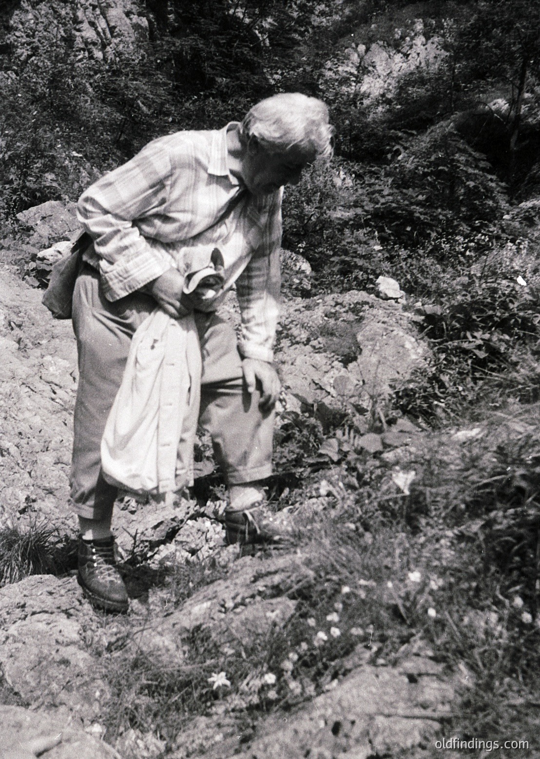 Mid-20th century hiker in rugged alpine terrain, examining a rock face. Lightweight hiking gear suggests mid-century mountaineering culture. Distinctive striped socks and rolled trousers hint at practical outdoor fashion.