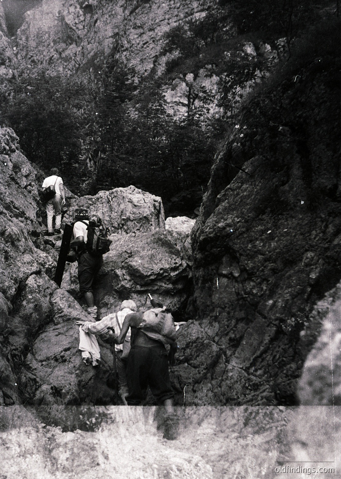 Mid-20th century black-and-white photo of three hikers navigating a rugged alpine trail with steep rock faces and misty waterfall. Clothing suggests 1950s–1960s outdoor gear. Dramatic lighting highlights texture and scale of mountainous terrain.