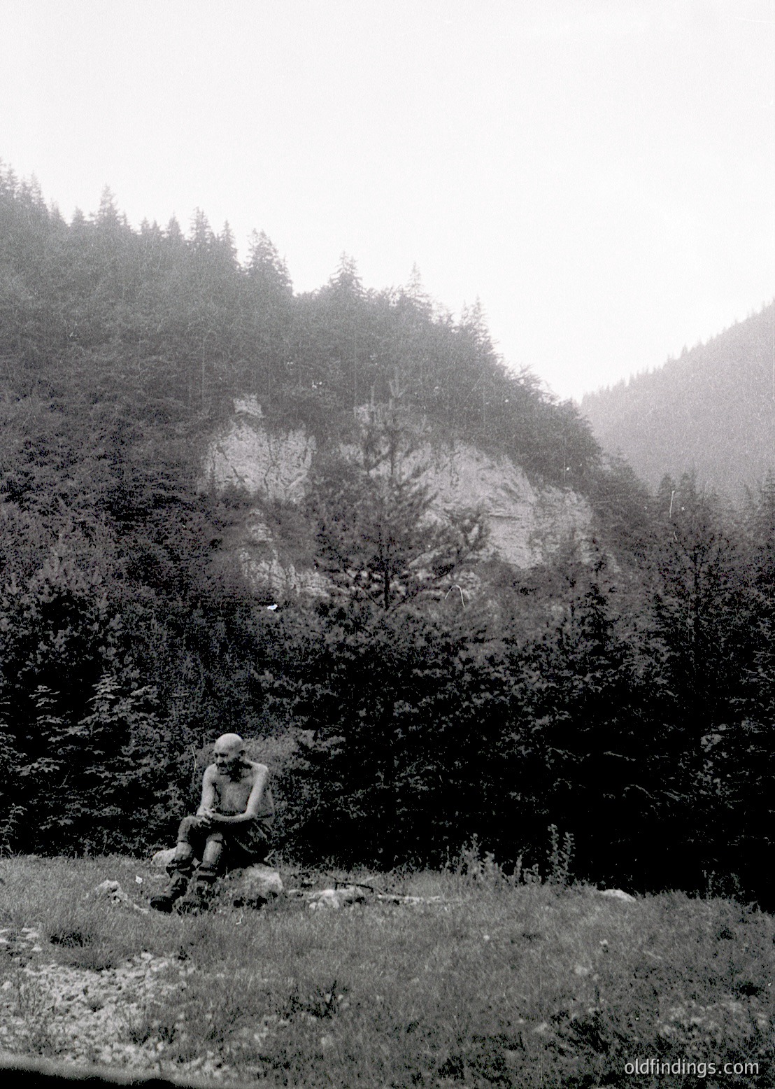 Black-and-white mountain scene featuring a lone figure in mid-hike, seated on a rock with backpack. Dense forest and misty slopes dominate the background, suggesting alpine terrain. Clothing and gear suggest mid-20th century outdoor recreation.