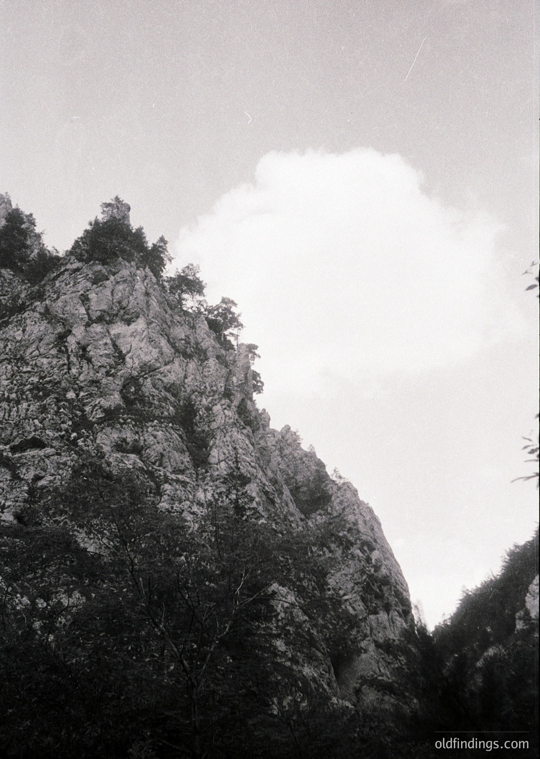 Monochrome mountain peak with rugged, vertical rock face and sparse vegetation. Dramatic cloud formation partially obscures summit, creating a striking silhouette. Likely captured in mid-20th century due to film grain and style.