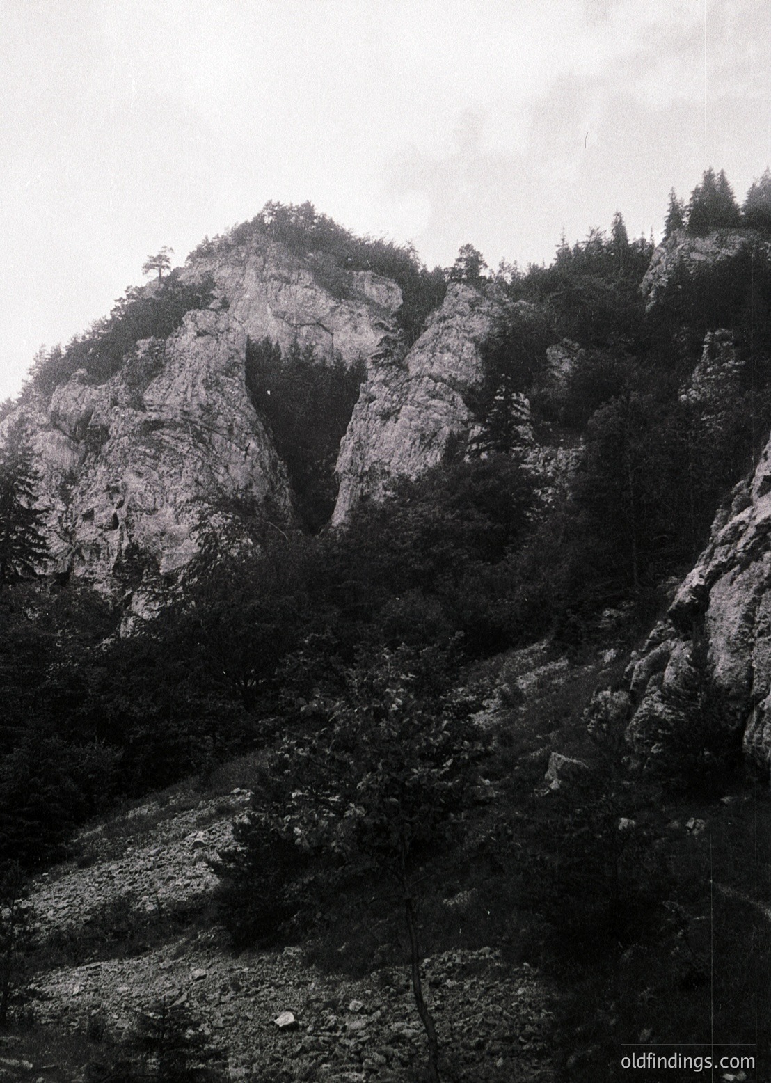 Monochrome mountain landscape with steep rocky slopes and sparse vegetation. Dramatic vertical rock formations dominate the scene, likely part of a rugged alpine or highland region. Fog or mist obscures the upper ridge, adding depth. Early-to-mid 20th-century vintage due to grainy texture and sepia tone.