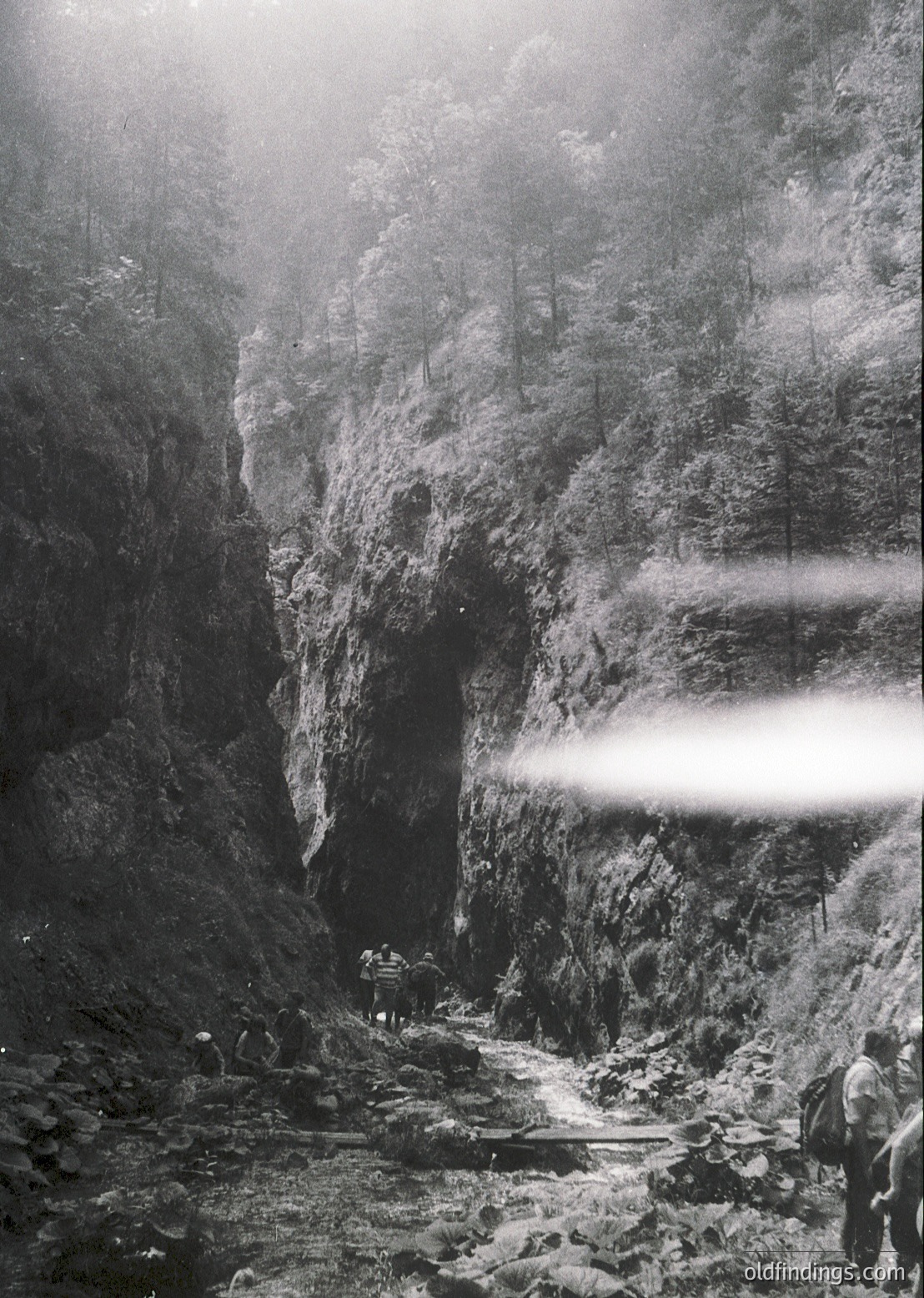 Sunlight filters through mist in this dramatic alpine canyon, illuminating rugged rock walls and a narrow, winding path. Three hikers in vintage gear (packs, long coats) navigate the rocky terrain, suggesting mid-20th century exploration. Fog obscures distant forest edges, enhancing the sense of isolation.