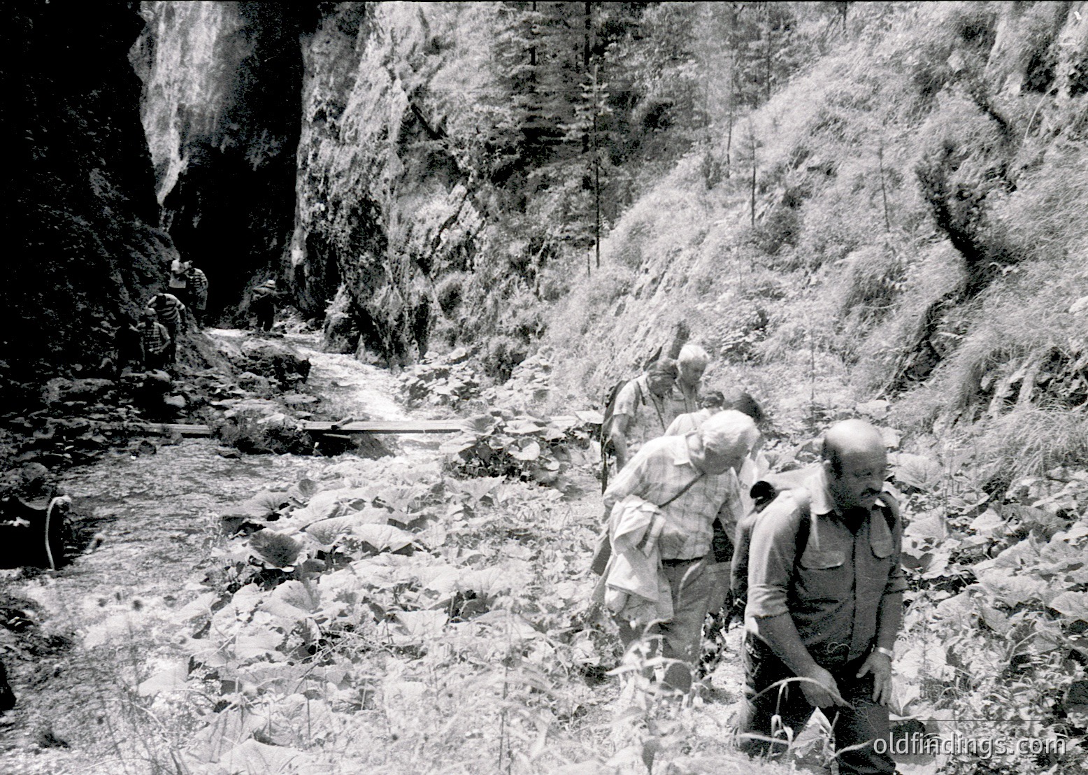 Three hikers in rugged alpine terrain, mid-20th century. Steep rock walls frame a narrow, rocky path with a trickling stream. One climber kneels, adjusting gear, while others stand or crouch. Classic mountaineering attire—wool caps, jackets, and sturdy boots—suggests a challenging ascent. Potential or region.