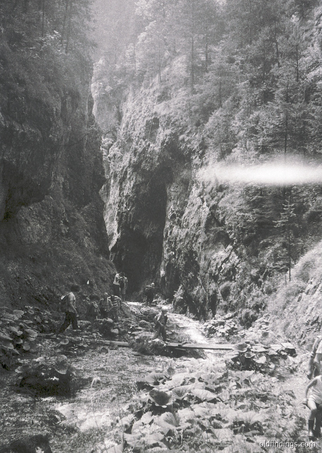 Black-and-white shot of a rugged mountain gorge with steep rock walls and a narrow, rocky path. Two hikers in mid-20th-century attire (long pants, boots) navigate the terrain, one carrying a backpack. Sunlight filters through mist, illuminating the path’s edge.