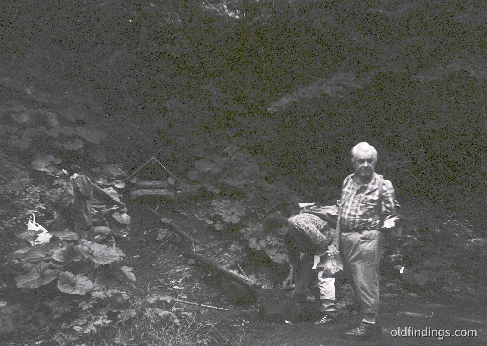 Black-and-white image of two men in rugged alpine terrain, likely mid-20th century. One man kneels near a small wooden structure, while the other stands with a dog, possibly surveying or preparing for a task. Snow patches and rocky ground suggest high-altitude or mountainous region. Clothing indicates outdoor work or exploration.