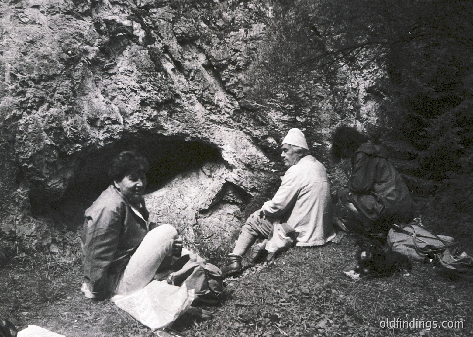 Three individuals examine a rock cave entrance, likely during archaeological excavation. Mid-20th century attire suggests or . One person kneels, holding tools, while others crouch or sit nearby. Natural rock formations dominate the background.