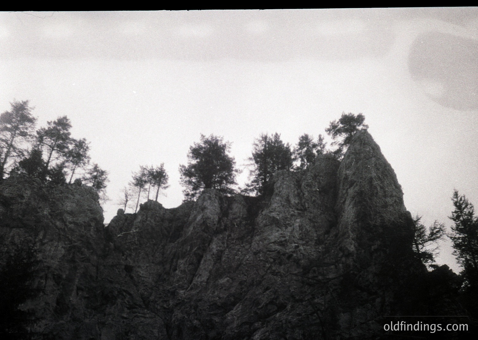 Monochrome landscape featuring jagged rock formations with sparse pine trees clinging to steep slopes. Dramatic, moody lighting enhances texture and depth. Likely captured in a high-altitude or alpine region.