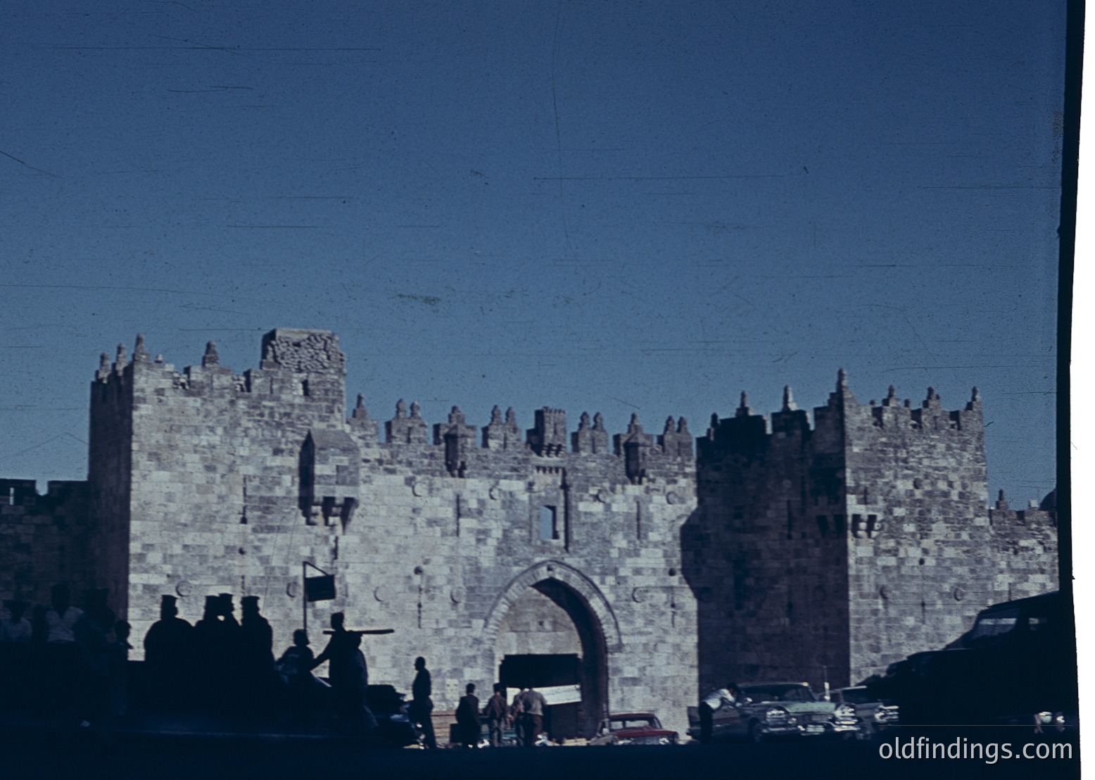 Historic stone gate with crenellated towers, likely the Jaffa Gate in Jerusalem’s Old City. Mid-20th century architecture with arched entrance flanked by battlements. Crowds and vintage cars suggest pre-digital era.