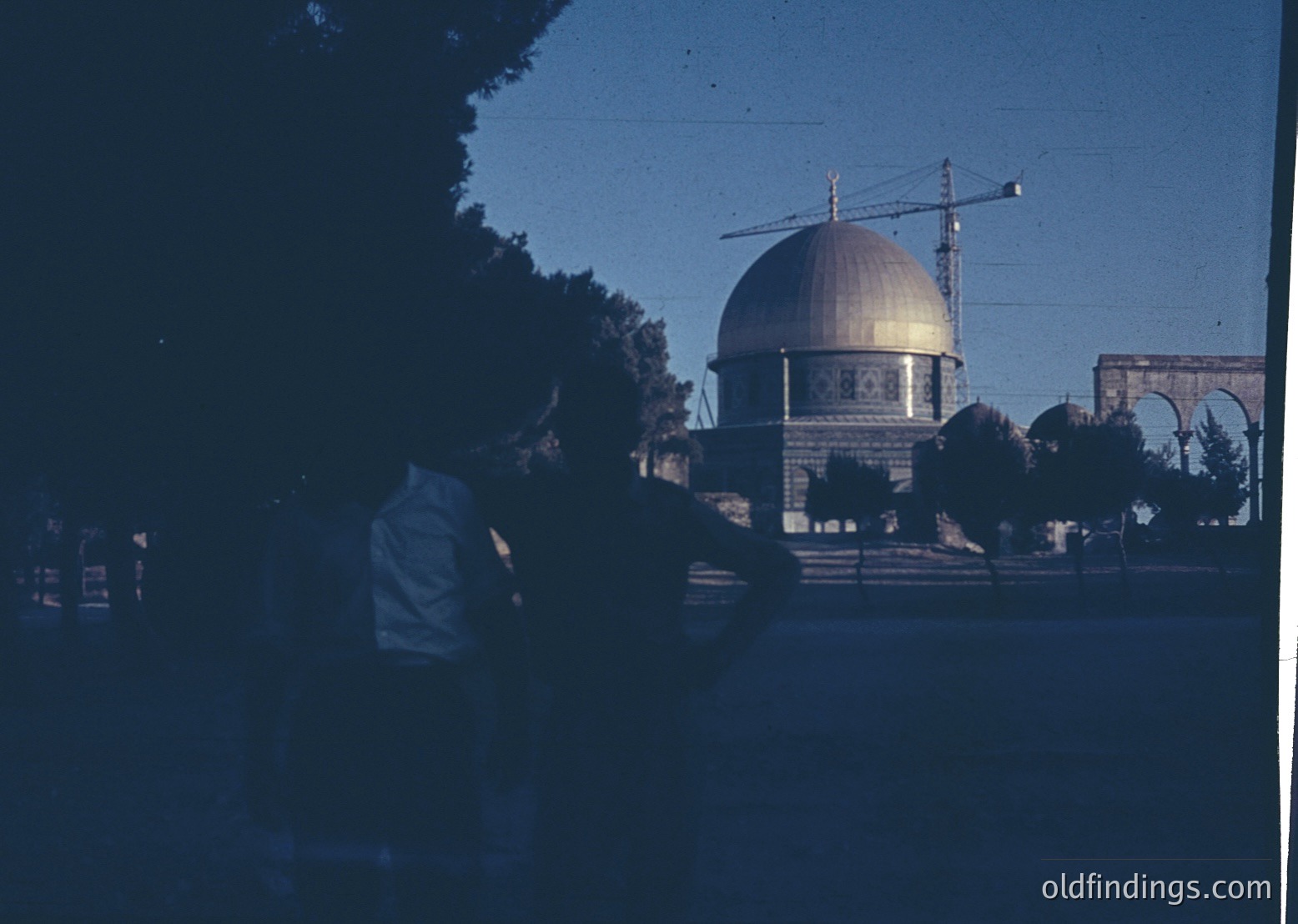 Vintage black-and-white photo of Dome of the Rock in Jerusalem, featuring its iconic golden dome and intricate geometric patterns. Construction crane in background suggests mid-20th-century urban development. Blurred figures in foreground add scale.