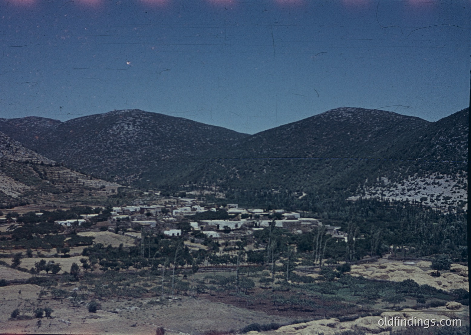 Vintage sepia-toned aerial view of a rural settlement nestled between barren hills, likely mid-20th century. Clustered low-rise buildings with flat roofs and sparse vegetation dominate the landscape. Power lines crisscross the area, indicating early electrification. The scene suggests a remote, agricultural community.