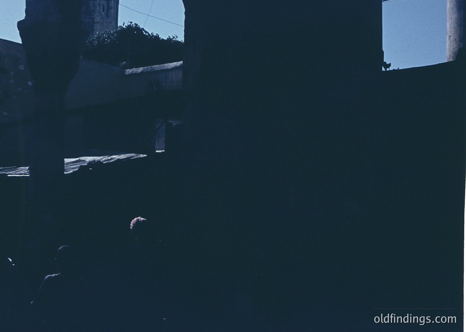 High-contrast monochrome shot of a shadowy interior space, likely a balcony or covered terrace. Partial view of a wooden railing and faint outline of a person sitting. Architectural details suggest mid-century modern design. Potential urban setting with visible greenery and distant structures.