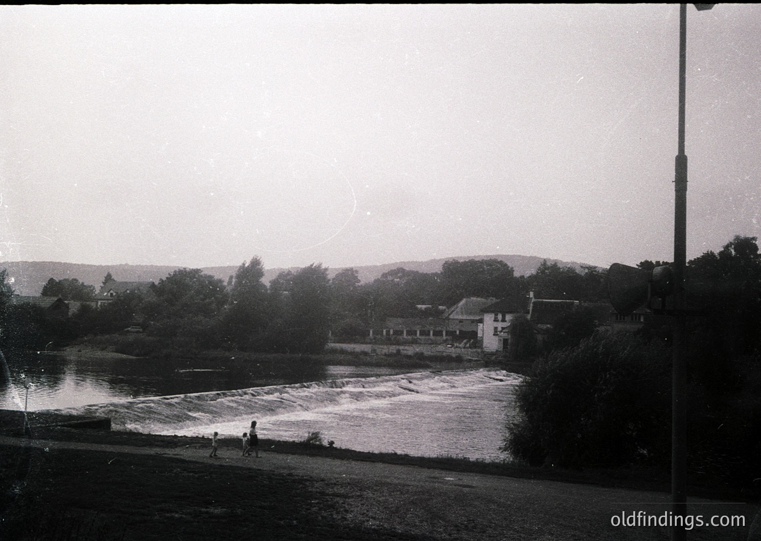 Black-and-white riverside scene with cascading waterfall, likely mid-20th century. Small group of people near water’s edge; distant buildings and trees frame serene landscape. Possible rural or small-town setting.