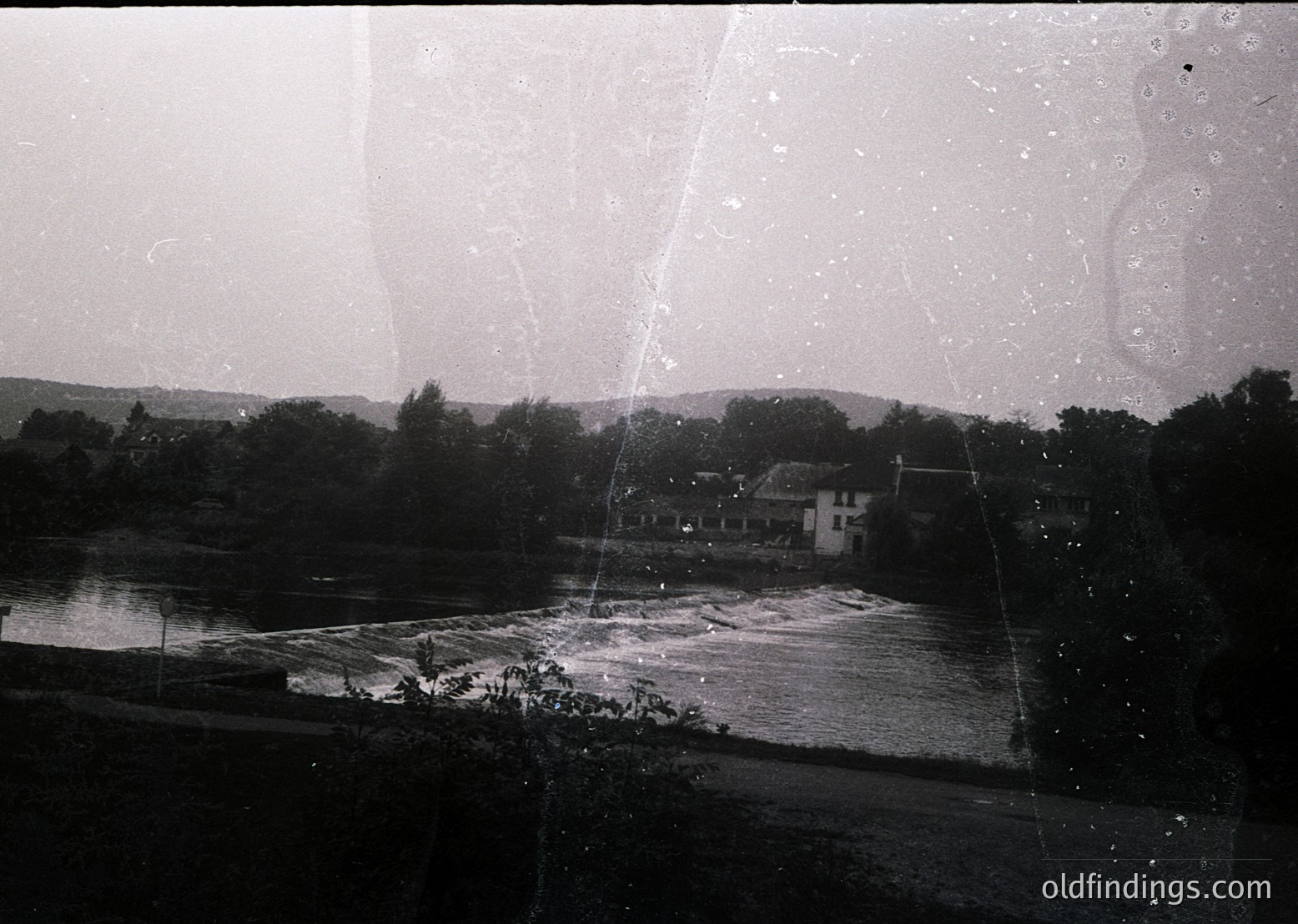 Vintage black-and-white photo of a rural riverside scene with a small cluster of buildings near water’s edge. Distinctive curved window frame in foreground suggests indoor perspective. Dense foliage and rolling hills in background. Likely mid-20th century European countryside.