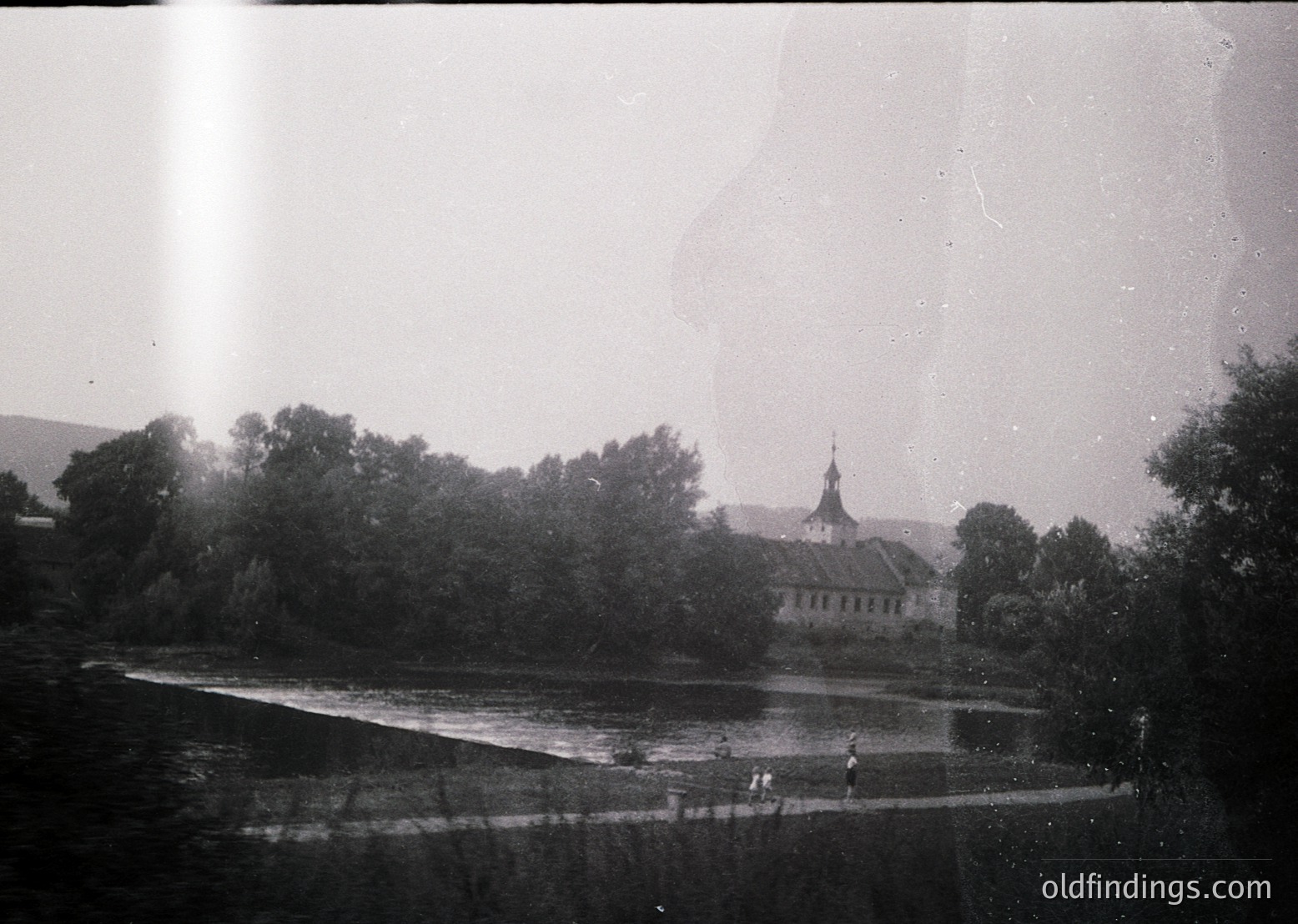 Vintage black-and-white photo of a riverside estate with classical architecture, likely mid-20th century. Prominent two-story building with a central tower and symmetrical façade, surrounded by dense greenery. Three figures in dark clothing wade in shallow water near the bank. Overcast sky enhances serene, timeless atmosphere.