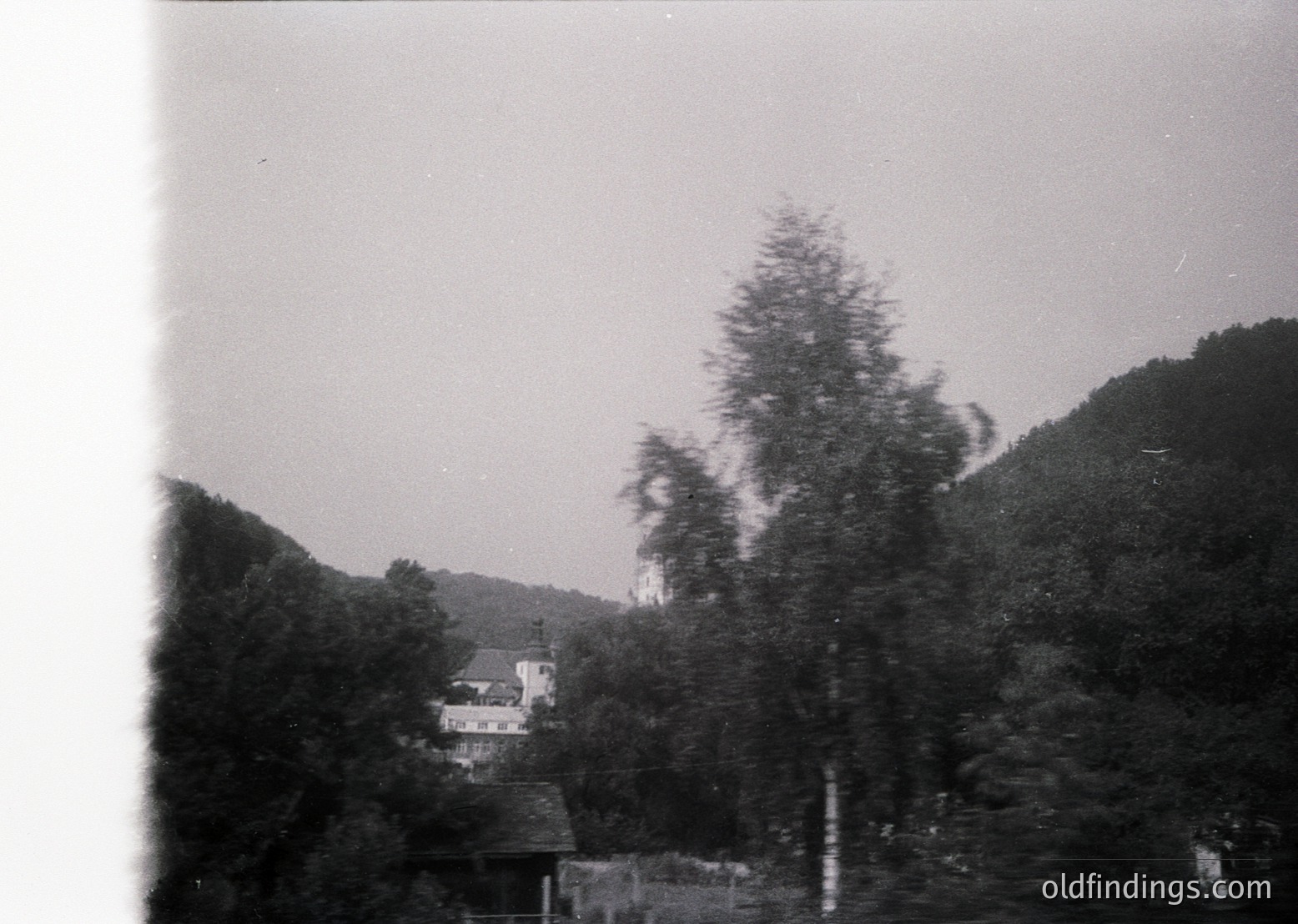 Vintage black-and-white rural scene featuring a modest wooden house nestled among dense forest. Sloped terrain and dense foliage dominate the background, suggesting a mountainous or hilly region. Likely mid-20th century due to architectural style and photographic grain.
