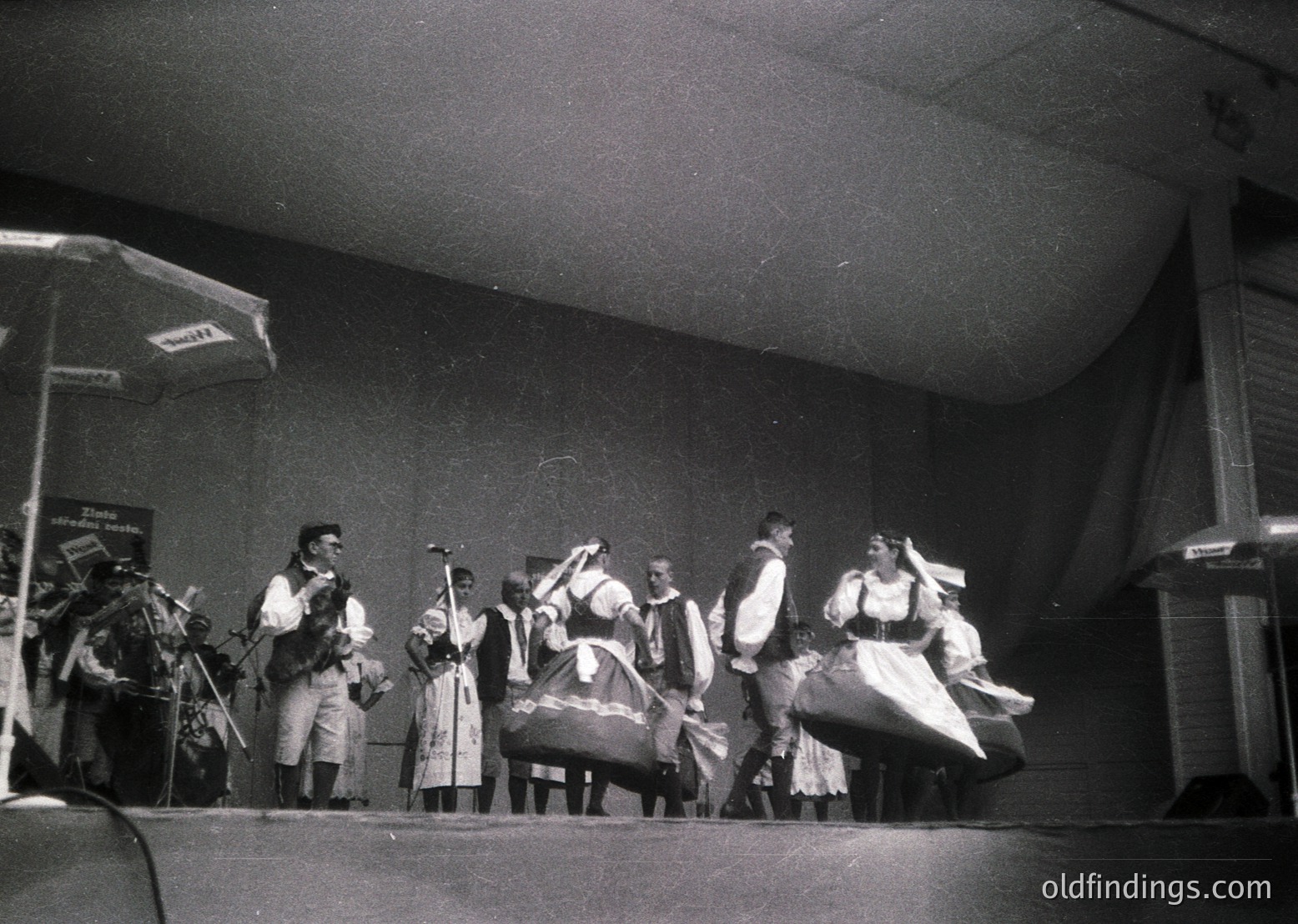 Traditional folk ensemble performing on stage, 1960s-1970s. Instruments include accordion, fiddle, and tambourine; women in embroidered dresses with shawls, men in vests and hats. Stage setup with microphone stand and banner.