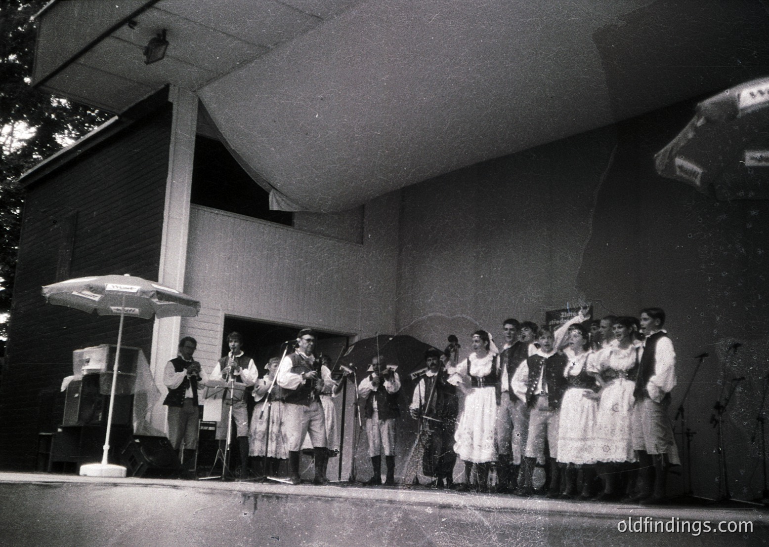 Black-and-white photo of a folk ensemble performing outdoors, likely 1960s–1980s. Traditional attire includes embroidered dresses, vests, and headscarves; instruments include accordions, violins, and drums. Stage setup features a small canopy and a speaker. Evokes Eastern European folk music revival.