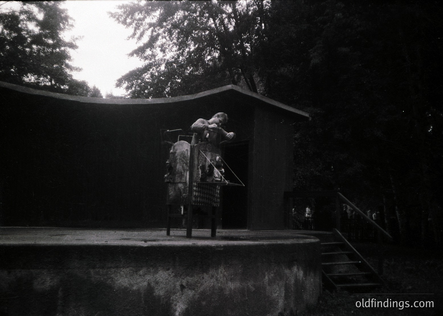 Black-and-white shot of a child on a wooden swing in a dimly lit park, framed by dense tree foliage. The swing’s metal frame and basket detail suggest mid-20th-century design. Concrete steps and a small building in background hint at public recreational space.