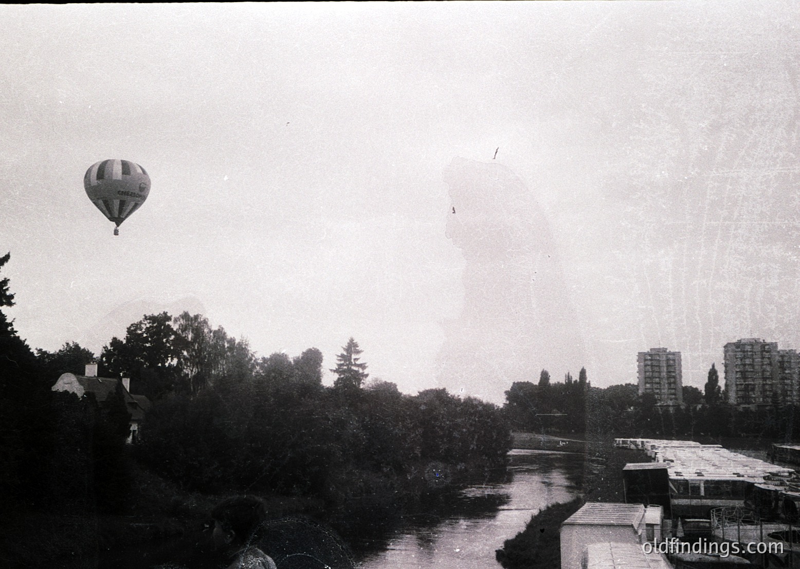 Mid-20th century urban scene with a hot air balloon over a river. Low-rise buildings and greenery frame the waterway, while high-rise apartment blocks loom in the background. Vintage black-and-white film captures industrial-era cityscape.