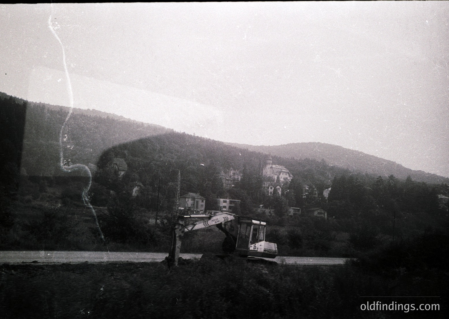 Black-and-white shot of a backhoe loader excavating on a rural roadside, framed by a window reflection. Dense forest and rolling hills in background suggest a mountainous region. Mid-20th century construction activity, likely 1950s–1970s.