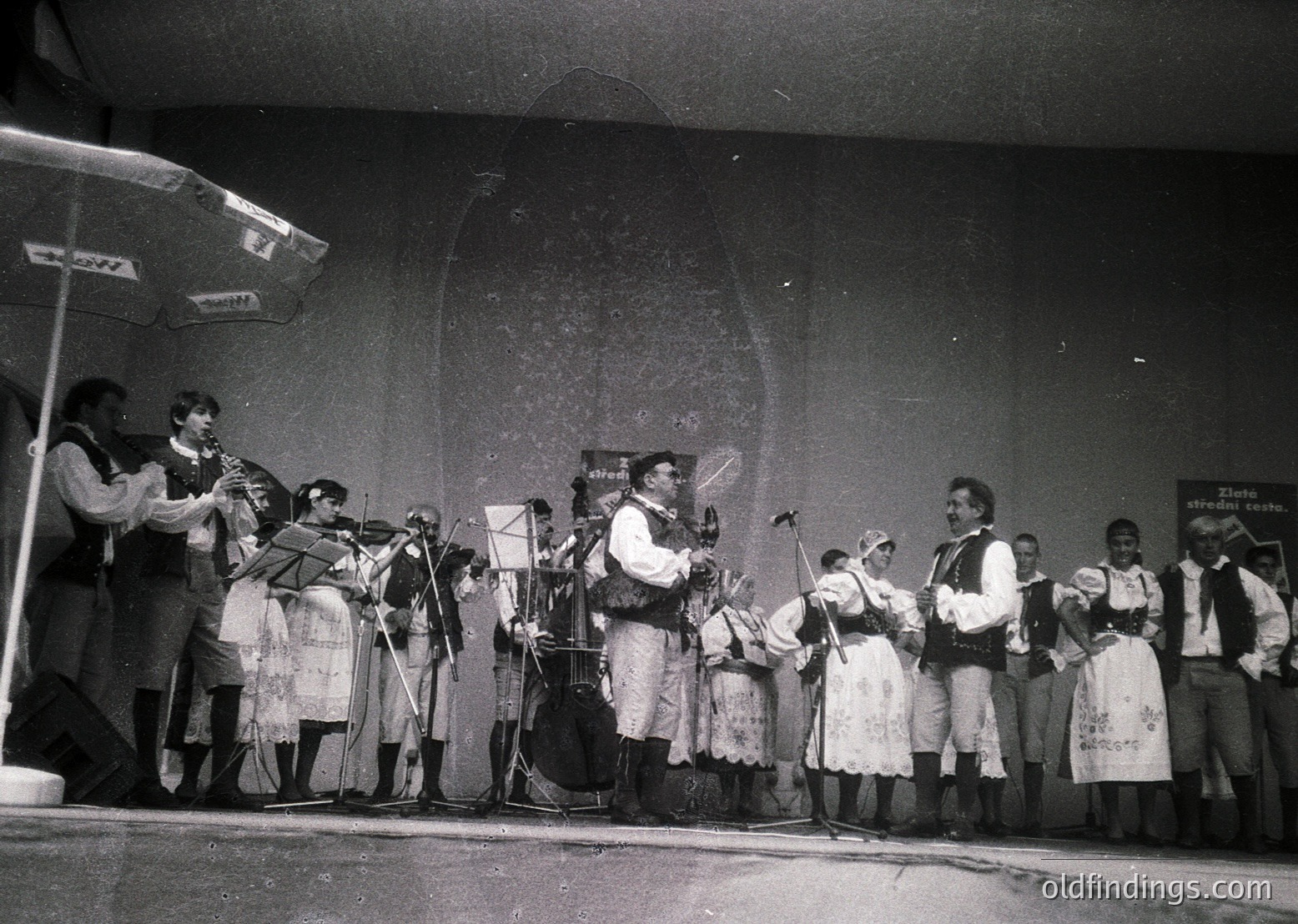 Traditional folk ensemble performing on stage, likely Eastern European, mid-20th century. Instruments include accordions, violins, and a drum. Participants wear embroidered folk costumes with aprons and vests. Stage backdrop features faded text ("Zlatni otredi" visible). Black-and-white, suggesting 1950s–1970s.