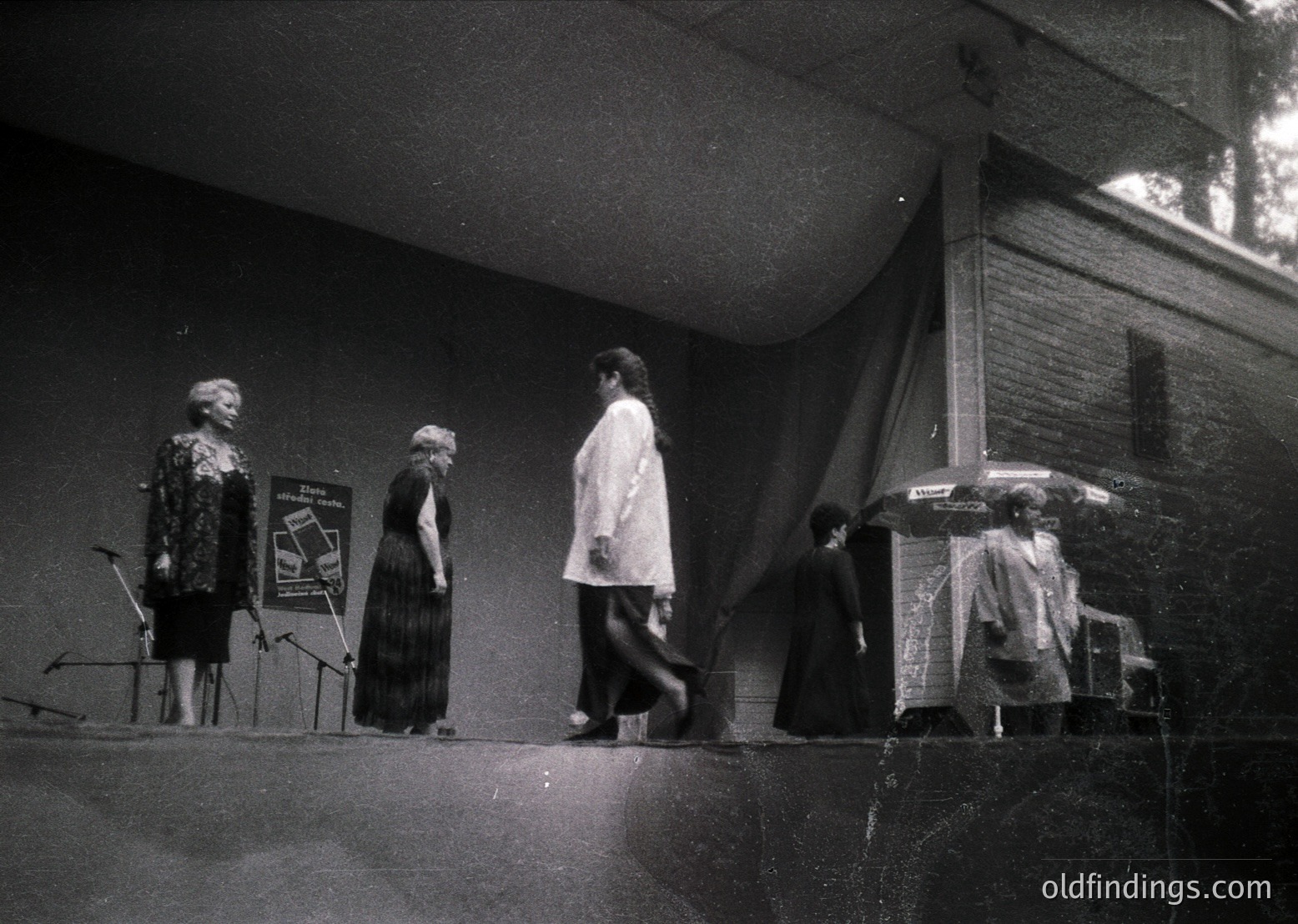 Black-and-white stage scene featuring three women in mid-20th-century attire: one playing piano, another standing beside a sign, and a third descending stairs. Stage lighting highlights their silhouettes against a dark backdrop. Possible concert or performance setting, 1950s–1960s.